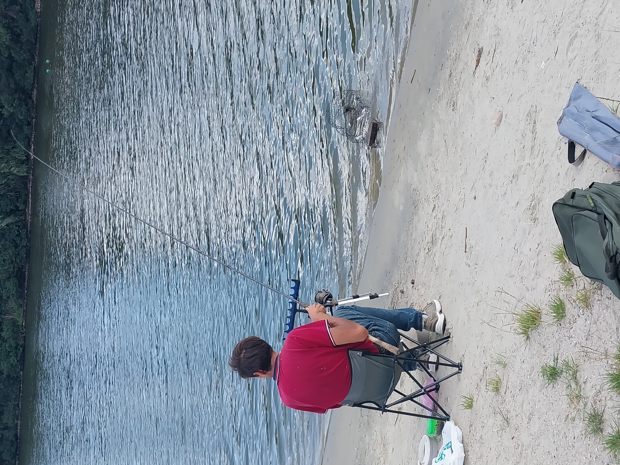 The image depicts a person fishing on the sandy bank of a body of water, likely a river or lake, with a dense line of trees in the background. The person is seated on a portable folding chair, wearing a red shirt and blue jeans, and is holding a fishing rod. Equipment for fishing, including a rod holder, a tackle box, and a fishing net submerged in the water near the bank, is visible. The water is rippling, and two green buoys are floating on the surface in the distance. The ground is sandy with sparse patches of grass, and two bags are placed on the sand near the fisherman. The scene appears to be outdoors in a natural setting.