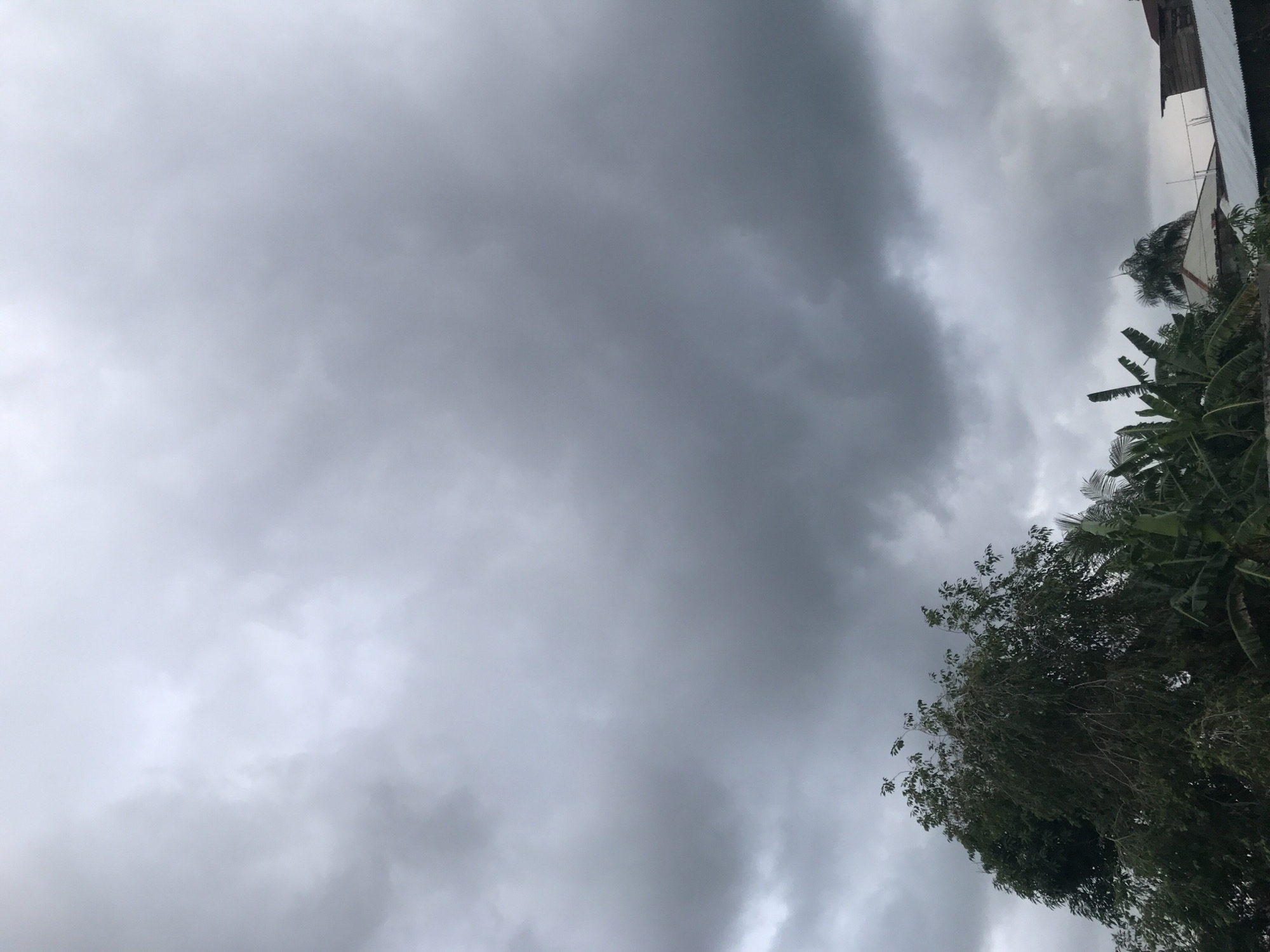 The image depicts a cloudy sky over Lhokseumawe, Indonesia. The sky is predominantly gray with varying shades of clouds. In the lower right corner, trees with green foliage are visible. A building structure is partially visible at the top right edge of the image.