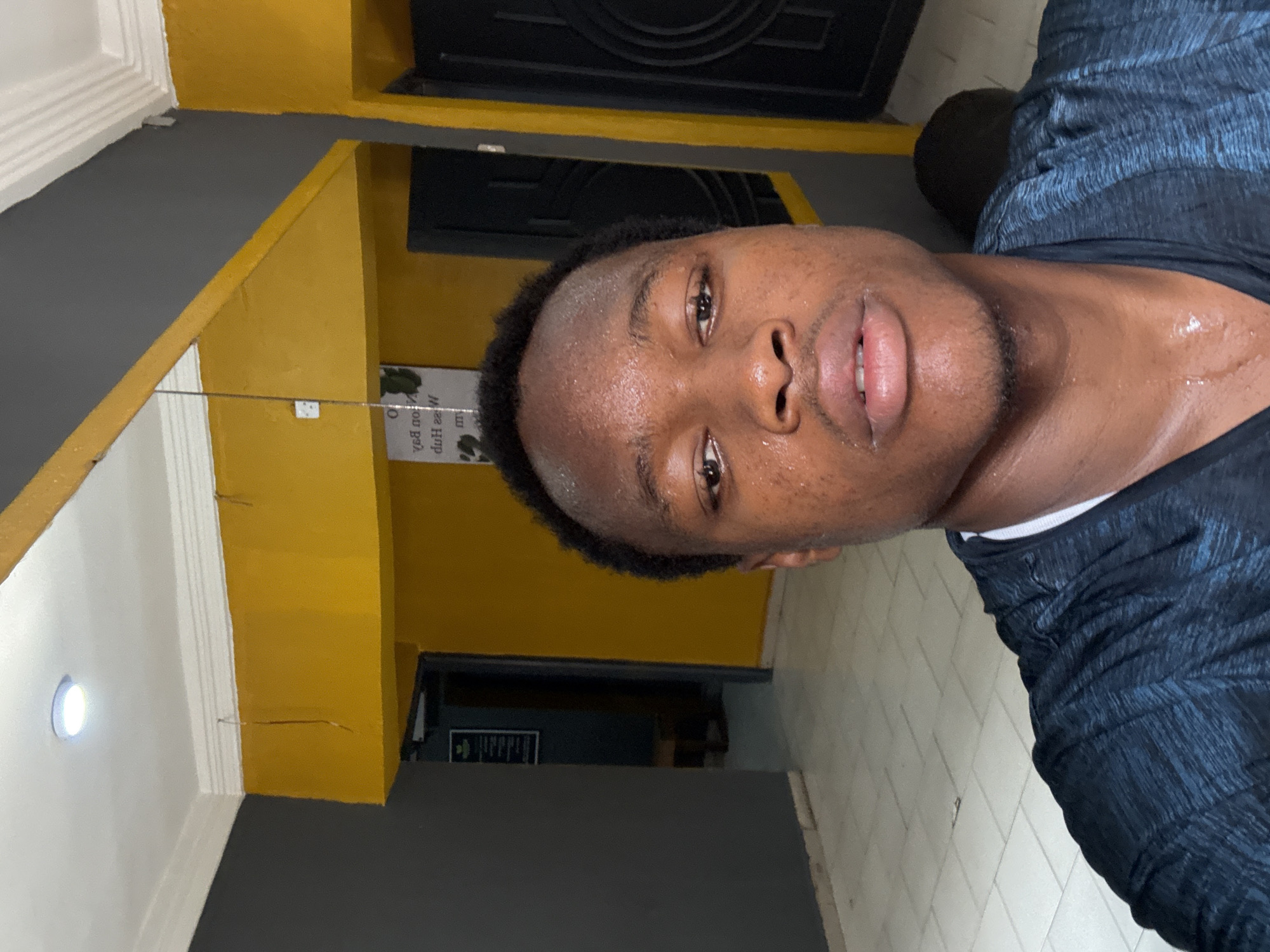 A young Black man is taking a selfie in what appears to be a gym or fitness center. He is visibly sweating, indicating he has likely just completed a workout. His face is moist, and sweat can be seen on his forehead, neck, and upper chest. He is wearing a dark blue, heathered t-shirt.
The background is a brightly lit indoor space with contrasting wall colors. A section of the wall is painted bright yellow, while another section is dark gray. The ceiling is white with molding. There is a light fixture on the ceiling and what looks like a mirror or reflective surface on the yellow wall, reflecting a white power outlet and a piece of string. To the left, a doorway leads to another area with a sign on the wall. To the right, another doorway or entrance is visible. The floor is tiled with a light-colored pattern.
The image does not contain any specific visual cues that definitively place it in Enugu, Nigeria.