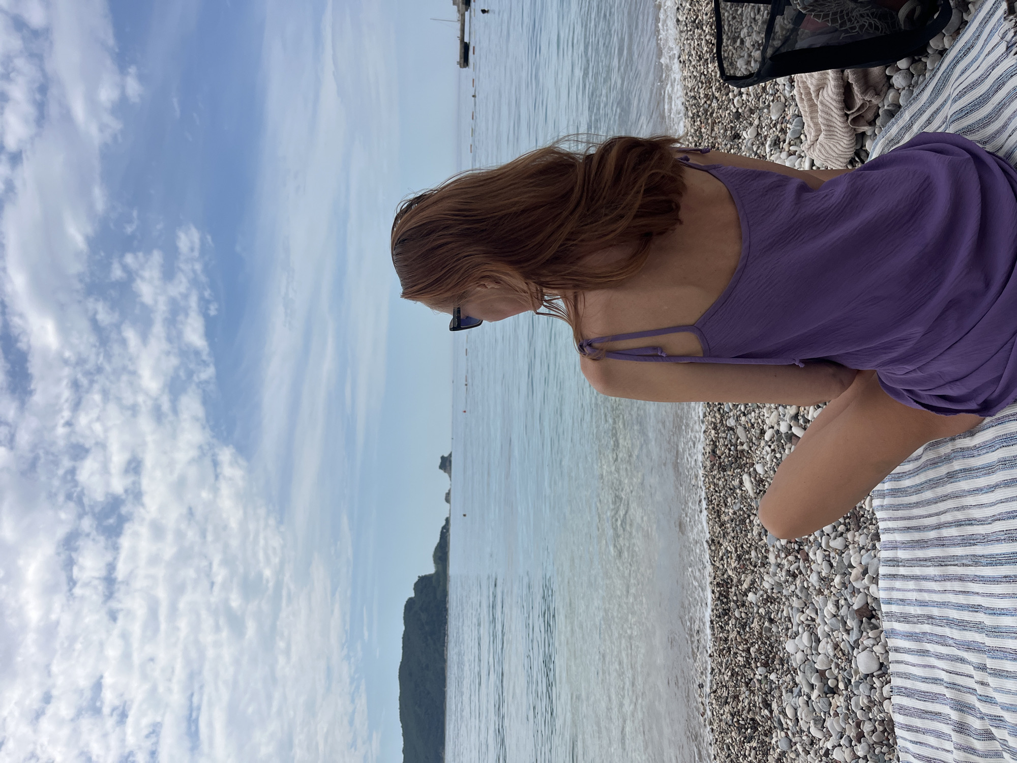 A medium shot of a fair-skinned woman with red hair sitting on a striped blanket on a rocky beach, gazing out at the sea in Budva, Montenegro. She is wearing a sleeveless, lavender-colored dress and sunglasses. The beach is composed of small, gray rocks. The water is clear and calm, with small waves lapping at the shore. In the distance, there is a green, rocky island and several boats. The sky is blue with scattered white clouds. A black mesh bag and a beige towel are partially visible in the upper right corner.