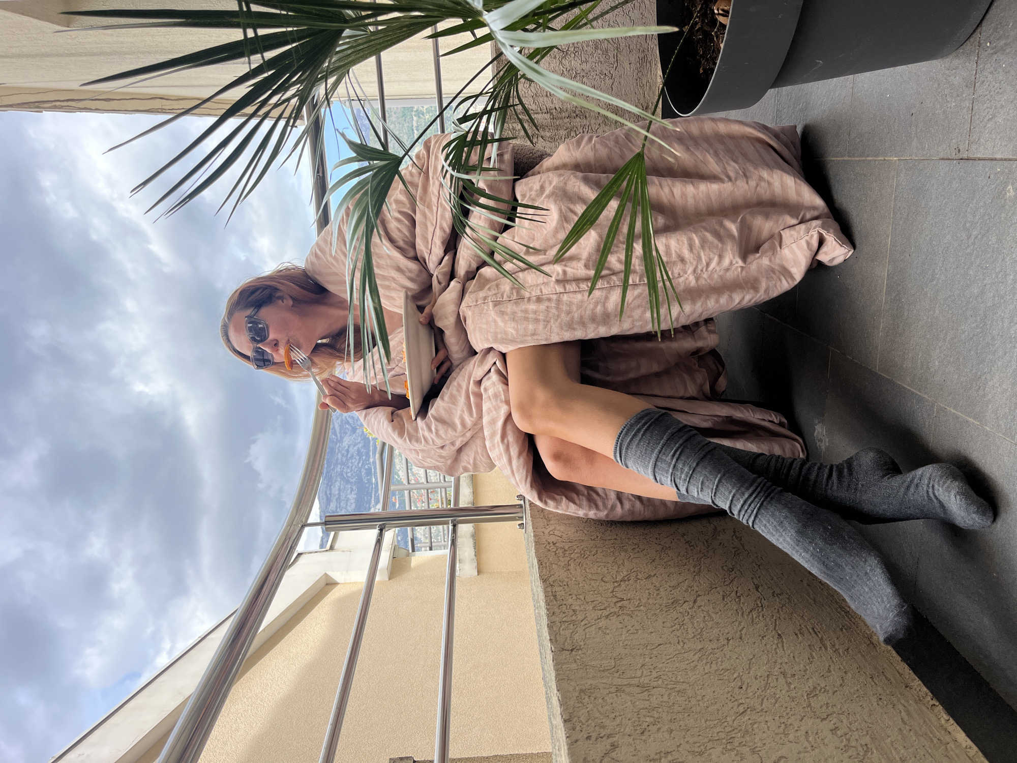 A woman is sitting on a balcony in what appears to be Budva, Montenegro. She is wrapped in a light pink striped blanket, wearing grey knee socks and sunglasses. She is eating something with a fork and holding a white plate. A plant is beside her in a black pot. The balcony has a metal railing and a textured wall. In the background, there is a cloudy sky and a view of a city and mountains.