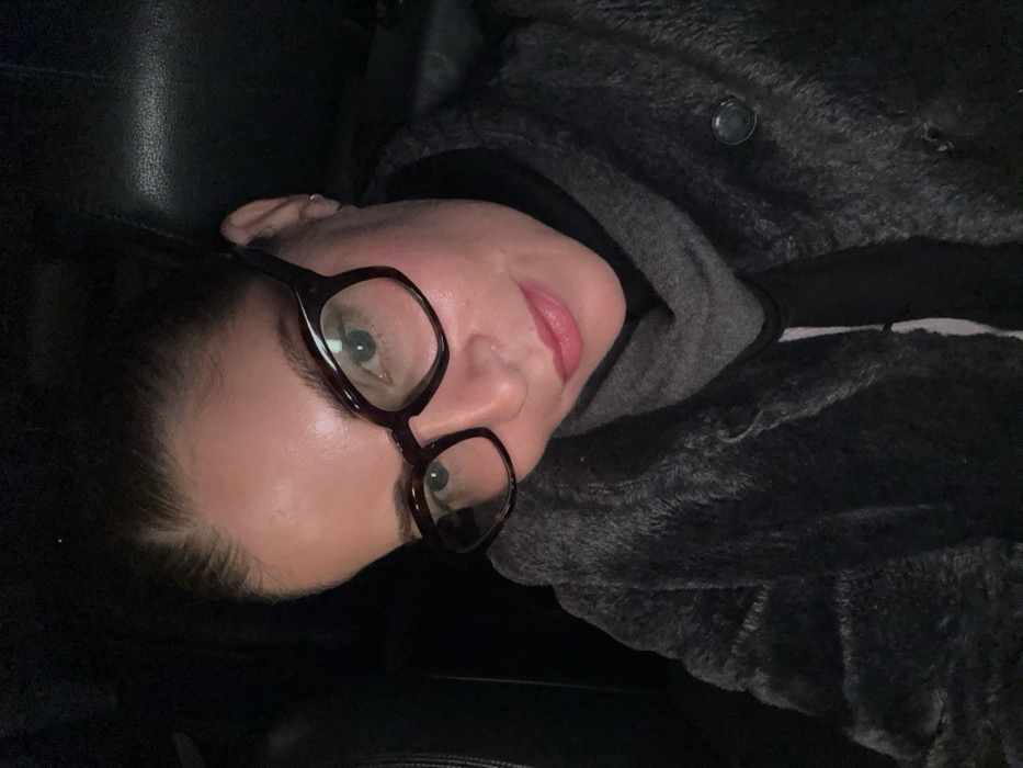 A woman with fair skin, wearing dark-rimmed glasses, gazes directly at the camera. She has dark hair pulled back, revealing a small earring. Her lips are tinted a soft pink. She's dressed in layers: a dark top, a gray hooded sweatshirt, and a dark, plush jacket with a button visible. The background appears dark, possibly inside a car. The image is angled, with the woman's head tilted to the left.
