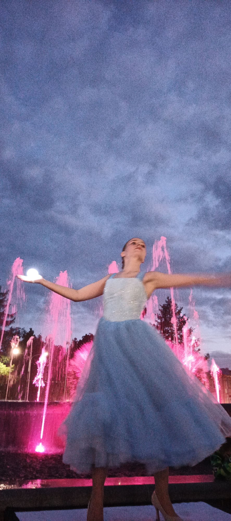 A young woman in a light blue, sparkly dress stands with her arms outstretched in front of a brightly lit pink fountain. She is holding a glowing orb in her left hand. The sky is dark and cloudy, suggesting it is twilight or night. The fountain is the main source of light, casting pink hues on the surroundings. The woman appears to be dancing or posing for a photograph. The background shows trees and some architectural elements, hinting at a park or public square setting.