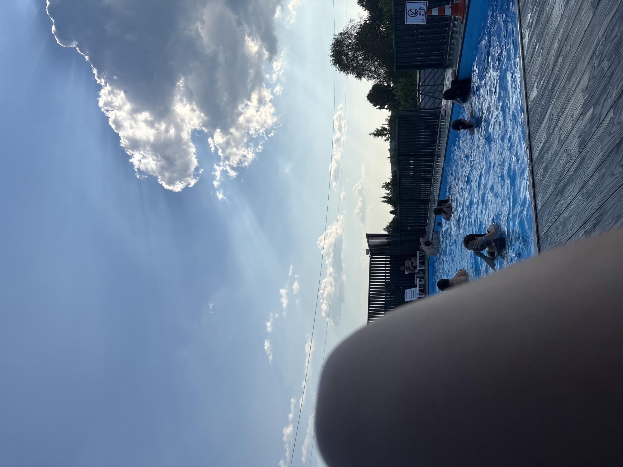 An outdoor swimming pool is pictured under a bright blue sky with sunbeams breaking through clouds. Several people are visible in the water, some swimming and others relaxing. The pool area is surrounded by a dark fence, and lounge chairs are placed on the deck. A sign with Russian text and a prohibition symbol is visible on the right side of the image. The foreground is partially obscured by a blurred, fleshy object, likely a leg. The overall scene suggests a recreational setting during the daytime.