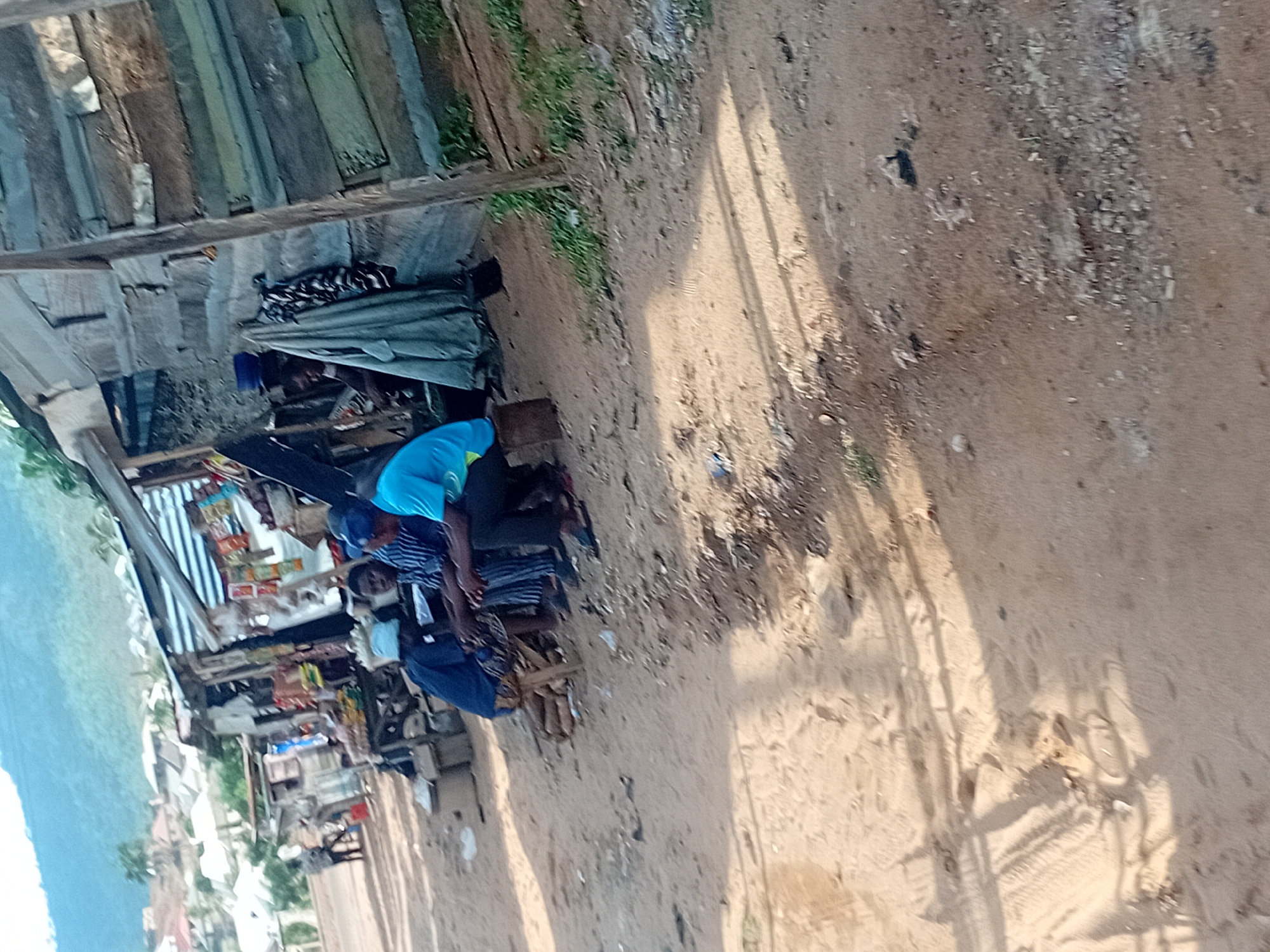 This media file depicts an outdoor scene, likely a market or roadside area in Lokoja, Nigeria. The ground is unpaved, consisting of sand and dirt with scattered small stones and debris. Long, distinct shadows are cast across the ground, suggesting it is either late morning or late afternoon under a clear sky.
In the mid-ground, a makeshift stall or small shop is visible. It is constructed from raw wooden planks and roofed with corrugated metal sheets. Inside the stall, shelves display various small packaged goods, possibly snacks, candies, or sachets of other items.
Several individuals are present near the stall. One person wears a bright blue t-shirt and dark trousers, seated on a low bench or stool. Next to them sits another individual in a striped shirt and dark trousers. Further to the left, closer to the stall's offerings, a third person is seated, clad in a blue outfit and a white head covering. The people appear to be engaged in casual interaction or waiting.
In the far background, a large, green mountain or hill range dominates the horizon, indicating a rural or semi-urban landscape. The overall scene portrays a common informal gathering and commercial point characteristic of such settings. No legible text is visible in the image.