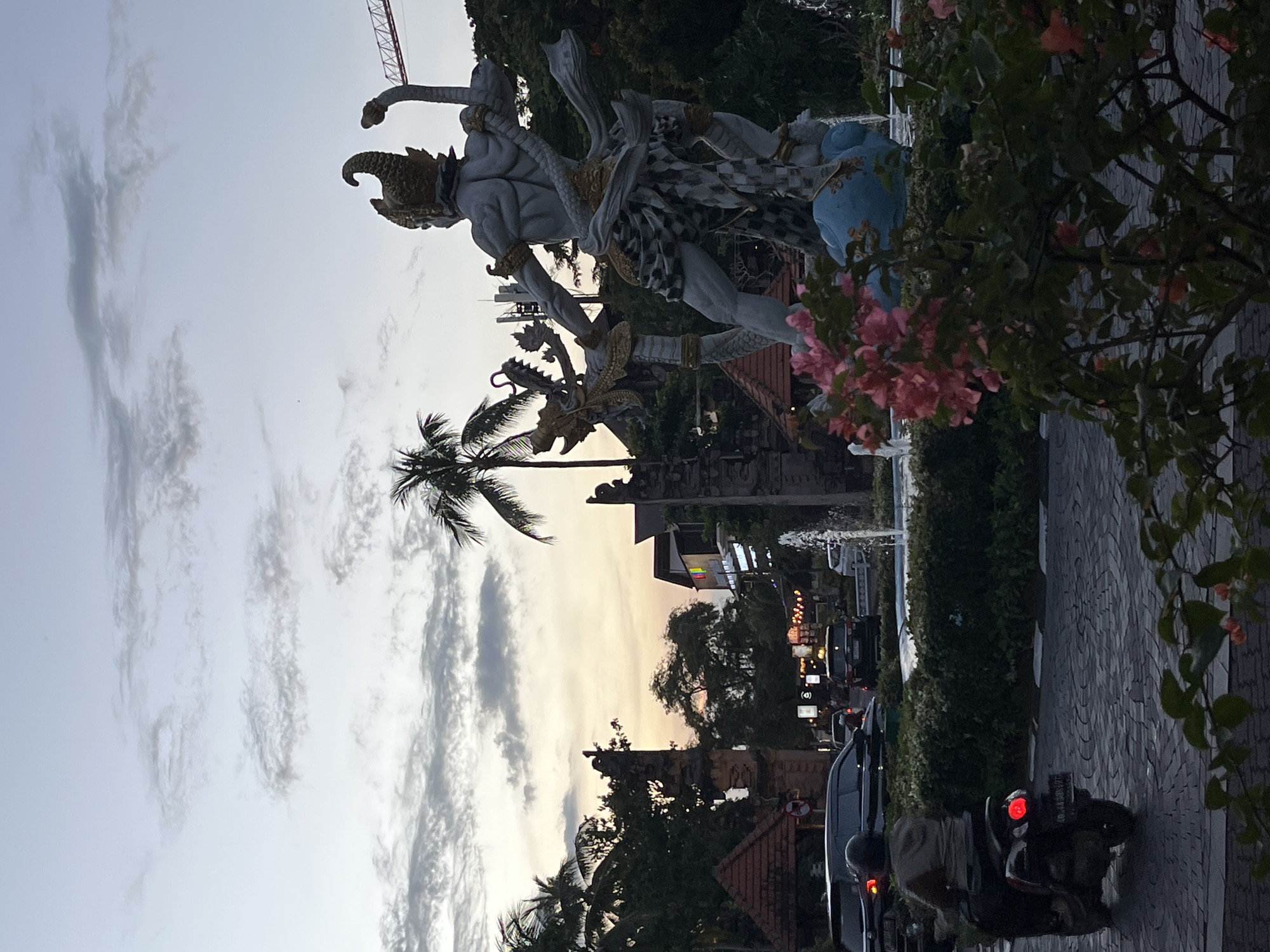 The image depicts a street scene in an urban area of Unknown, Indonesia, captured at dusk. In the foreground, a Balinese statue, possibly of a deity, stands prominently on a pedestal. The statue is a gray-white color with intricate details. To the right of the statue, a large flowering bush with pink blooms partially obscures the view.
The street is paved with gray bricks. Parked along the side of the road, a black car and a motorcycle are visible. A person, presumably the motorcyclist, is partially visible, wearing a helmet and neutral-colored clothing.
In the background, other buildings, trees, and streetlights are seen. The sky exhibits a mix of clouds and fading sunlight, suggesting the transition from day to night. A construction crane is also visible in the distance. A second statue is located at the center of the scene, partially concealed by palm fronds.