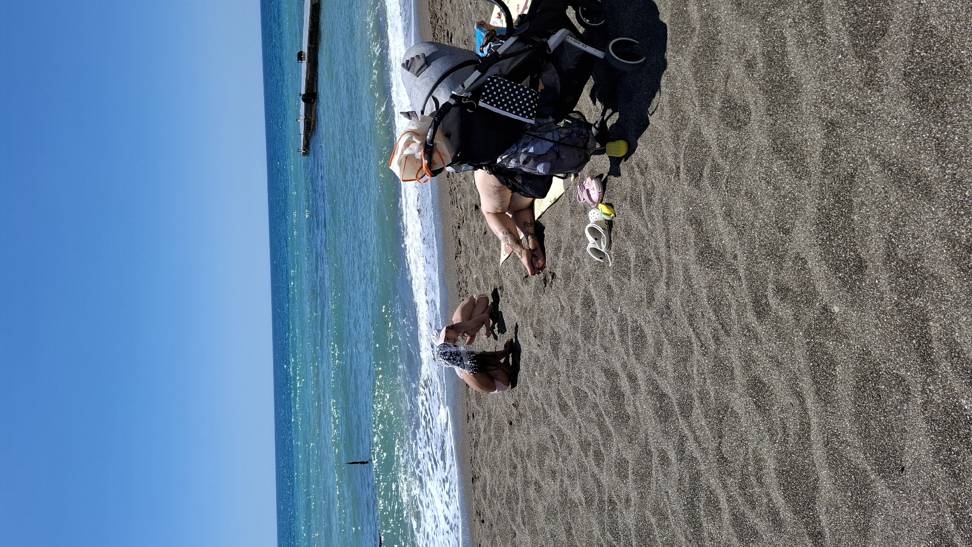 Here is a description of the image:
The image shows a sunny beach scene in an unknown location. The beach has dark sand and is adjacent to a clear, blue sea. In the foreground, a child wearing a patterned top and light-colored bottoms is playing near the water's edge. Nearby, an adult is kneeling or sitting on the sand. Next to the adult is a stroller. Several pairs of shoes are scattered on the sand near the adult. The sky is clear and blue. In the distance, there is a long pier or breakwater extending into the sea.