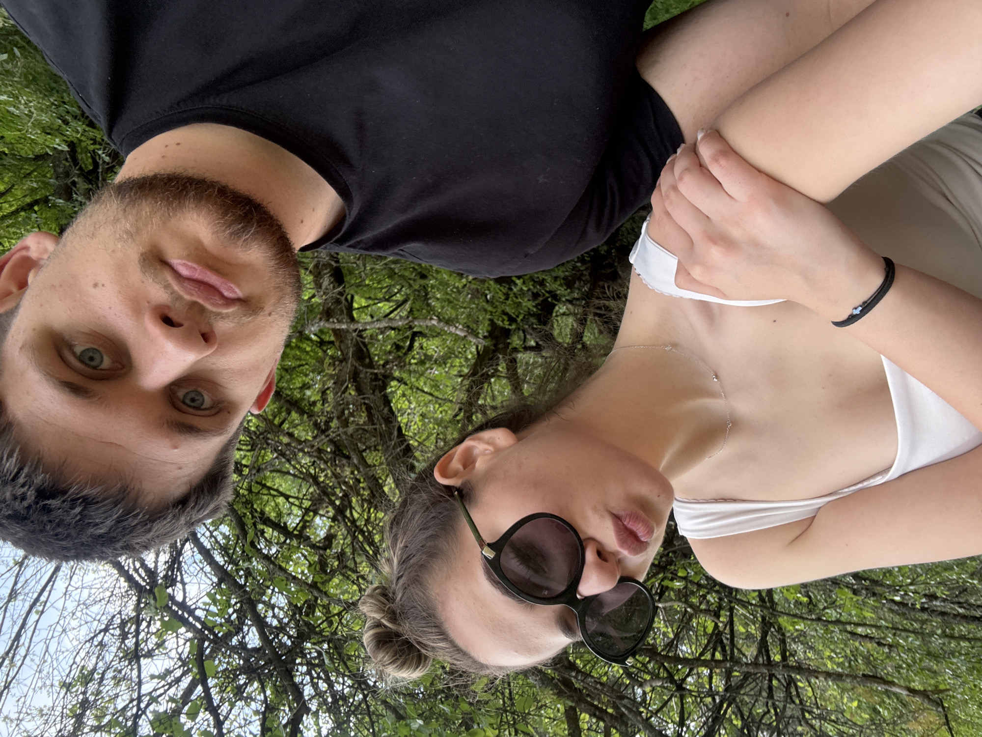 A selfie captures a man and a woman lying outside. The man, positioned on the left, has short gray hair, a light beard, and blue eyes. He is wearing a black T-shirt. The woman, on the right, has her hair pulled back in a bun and is wearing sunglasses and a white tank top. Their arms are intertwined. The background is filled with trees and foliage, suggesting a park or wooded area. Based on the details provided, the image could be taken in a park near the city of Blagoevgrad, Bulgaria.