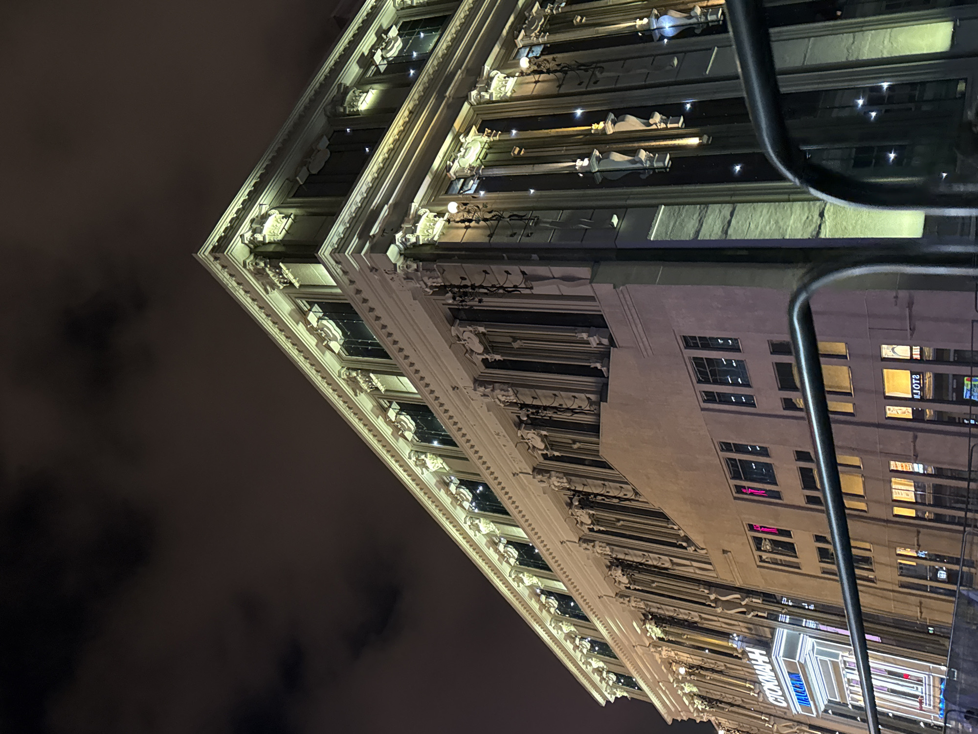 The image shows the TSUM department store in Yekaterinburg, Russia, at night. The building's facade is lit with warm yellow lights, highlighting its classical architectural details and rows of windows. The sky above is dark and cloudy. A black metal railing is visible in the foreground, suggesting the photo was taken from a slightly elevated vantage point, possibly a bridge or elevated walkway. Reflections of lights can be seen in the windows of the building.