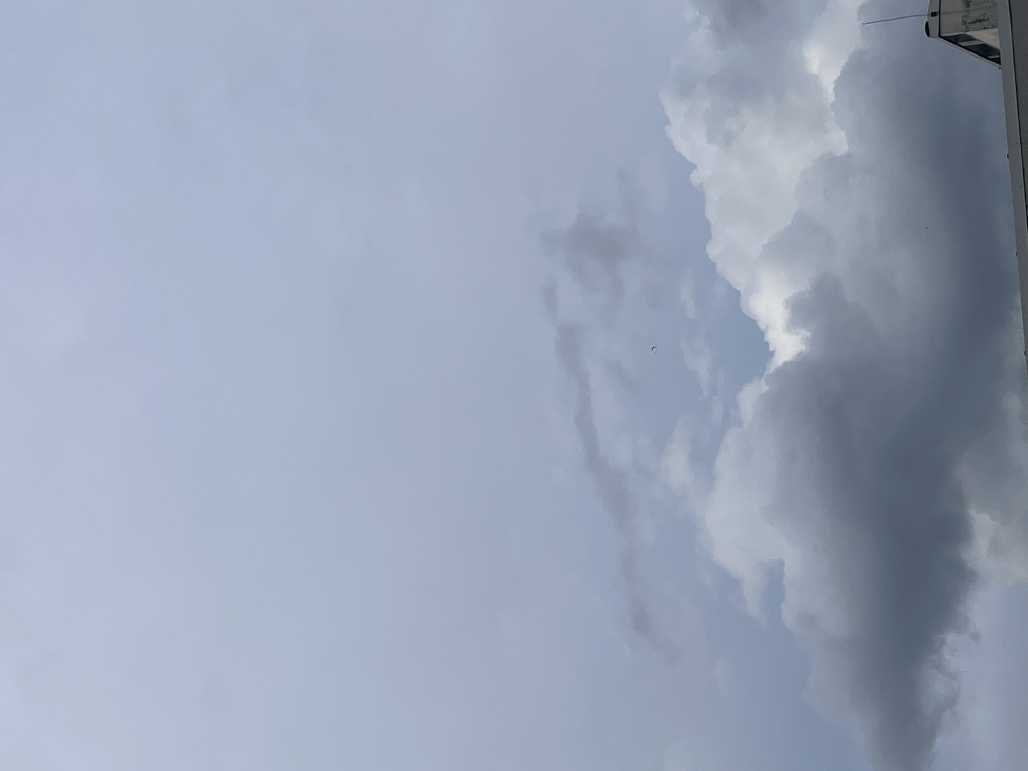The image shows a view looking up at a sky mostly filled with pale blue. Along the right edge are large, dark grey clouds. Part of a light-colored building is visible in the upper right corner. There is no visible evidence of a location in the city of Unknown, Unknown, people, objects, actions, or interactions.