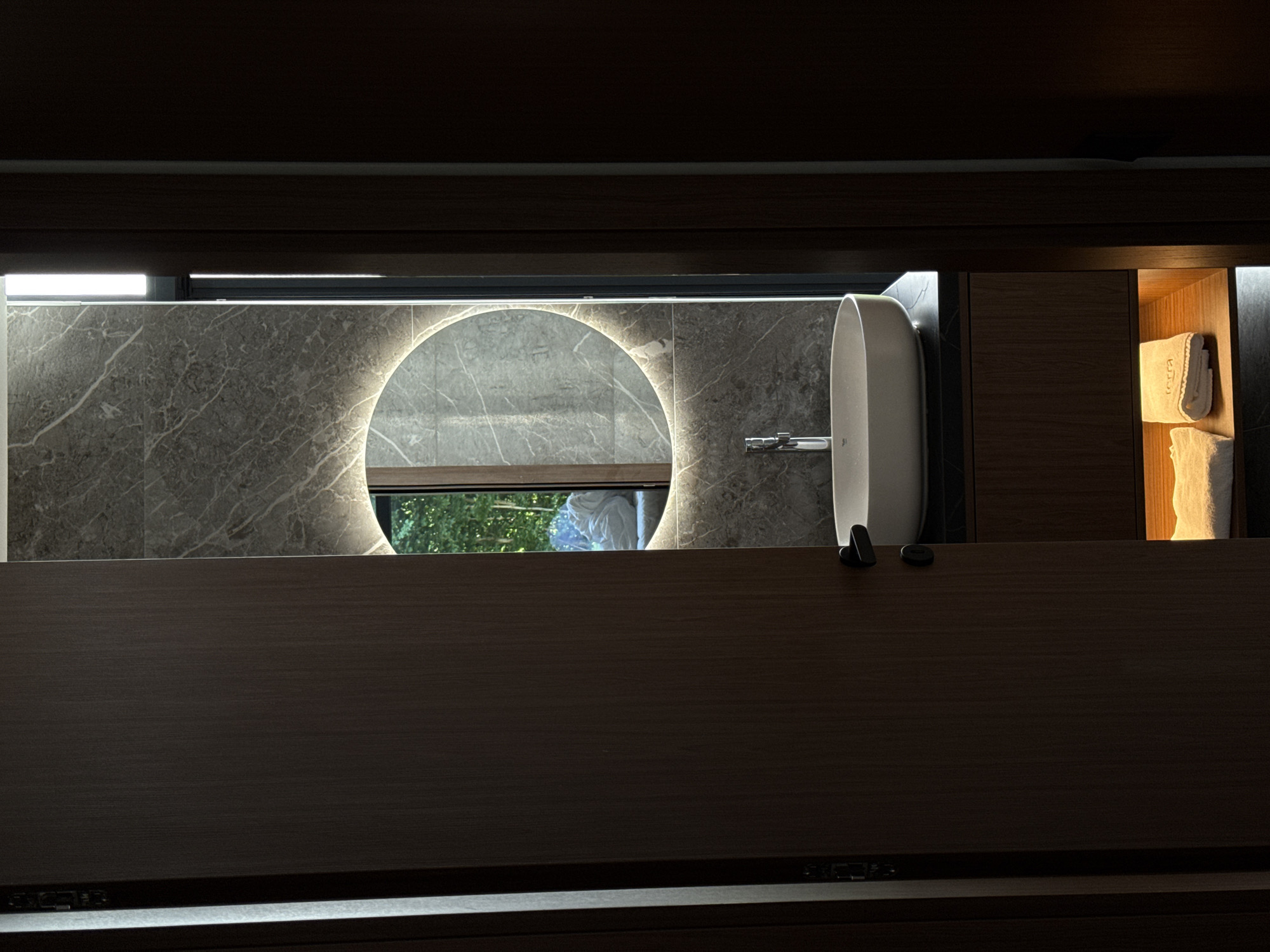 The image depicts a modern bathroom with a minimalist design, located in Bakovići, Montenegro. A dark wooden door is partially open, revealing a glimpse into the room. Inside, a sleek white countertop with a vessel sink is visible, complemented by a chrome faucet. Above the sink, a circular mirror with integrated LED lighting creates a halo effect. The wall behind the sink is clad in large-format grey marble tiles, adding a touch of luxury. A tall, thin window or light fixture runs vertically along the right edge of the tiled wall, allowing a sliver of natural light to enter. Below the countertop, a wooden cabinet houses neatly folded white towels illuminated by a soft, warm light from an internal shelf. In the reflection of the mirror, greenery and what appears to be a light-colored comforter or draped fabric can be seen, suggesting an outdoor view or an adjacent living space. The overall ambiance is one of sophisticated tranquility.