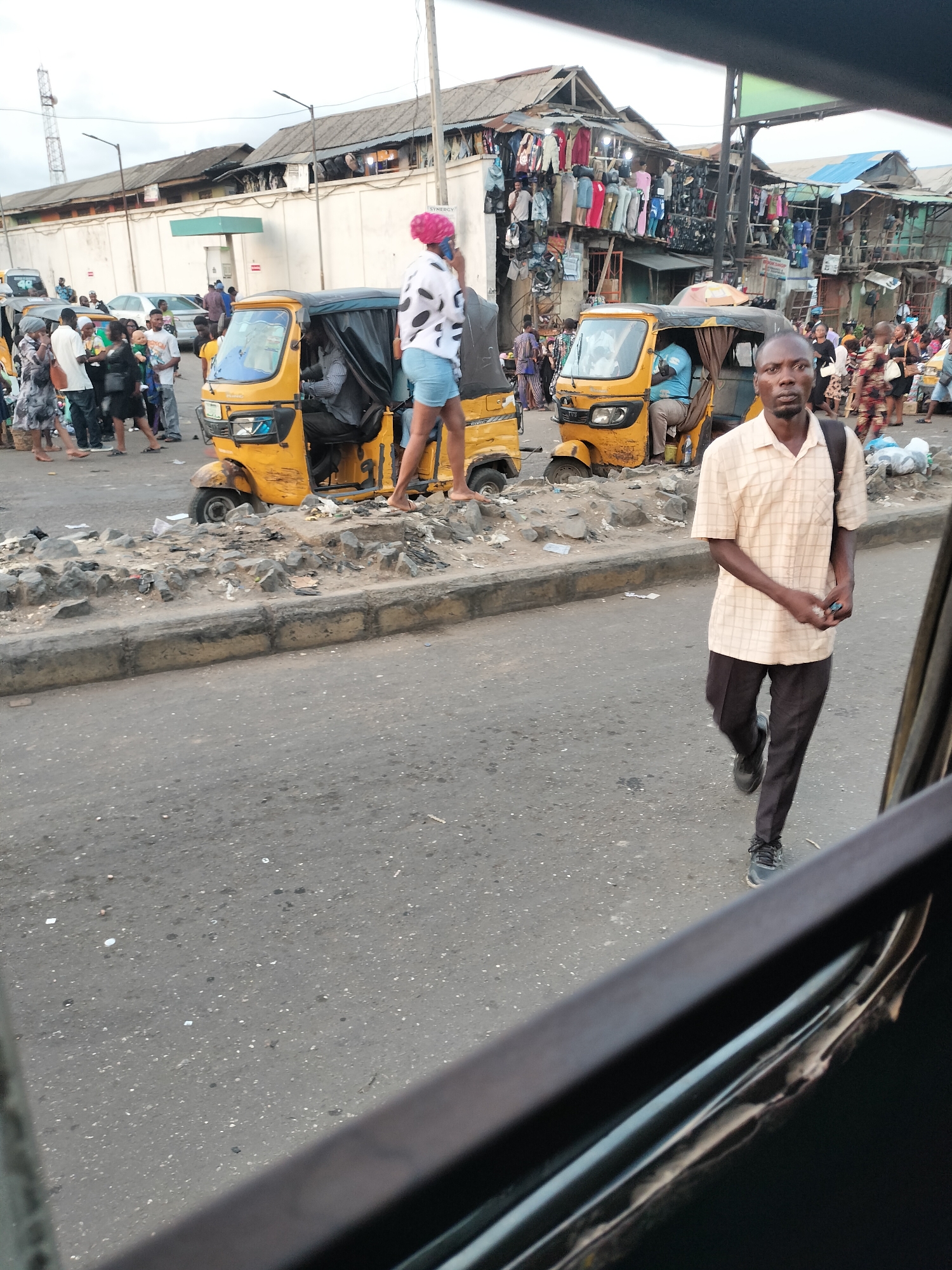 The image depicts a busy street scene in Lagos, Nigeria. Several people are visible, including a man walking in the foreground. He is wearing a checkered shirt, dark pants, and sneakers. Behind him are two yellow auto rickshaws (tuk-tuks) stopped on the side of the road. One woman with a pink head covering and light-colored clothing is standing on one of the vehicles. A crowd of people is visible on the left side of the image, near a white wall with "SYNERGY" printed on it. The background features a chaotic marketplace with clothing and various items for sale. The image is taken from inside a vehicle, with part of the window frame visible in the foreground.