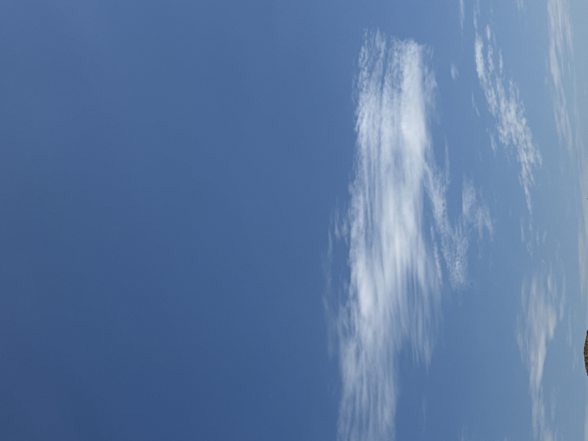 A vertical, medium-shot photo shows a bright blue sky with a long, thin, wispy cloud stretching horizontally across the middle of the frame. The cloud appears to be composed of cirrus formations, indicating fair weather. The bottom edge of the image contains a small, dark, textured area that appears to be the top of a building or structure, suggesting it is a terrestrial scene. The location is Ostrówek, Poland.