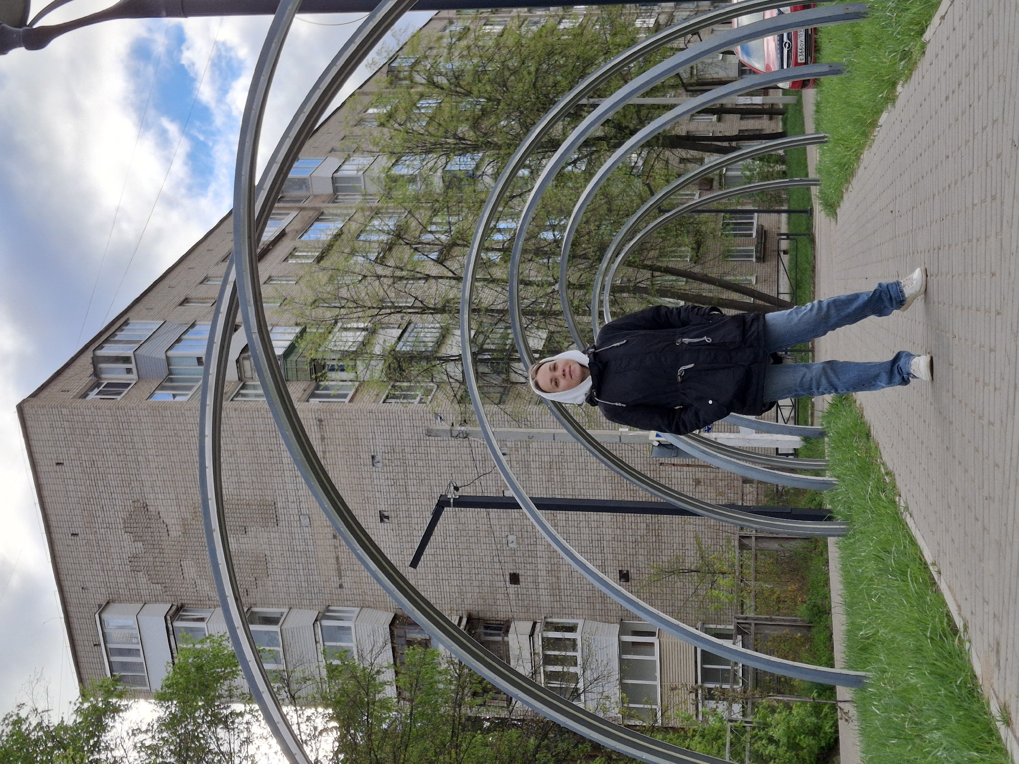 A person wearing a black jacket and blue jeans is standing under an array of metal arches in Chaikovsky, Russia. The person is facing the camera and is slightly turned to the right. A paved walkway runs alongside the metal structure with grass growing along the edge. In the background is a tall brick apartment building with several windows and balconies. The sky above has white clouds and patches of blue. A red car is partially visible behind one of the arches.