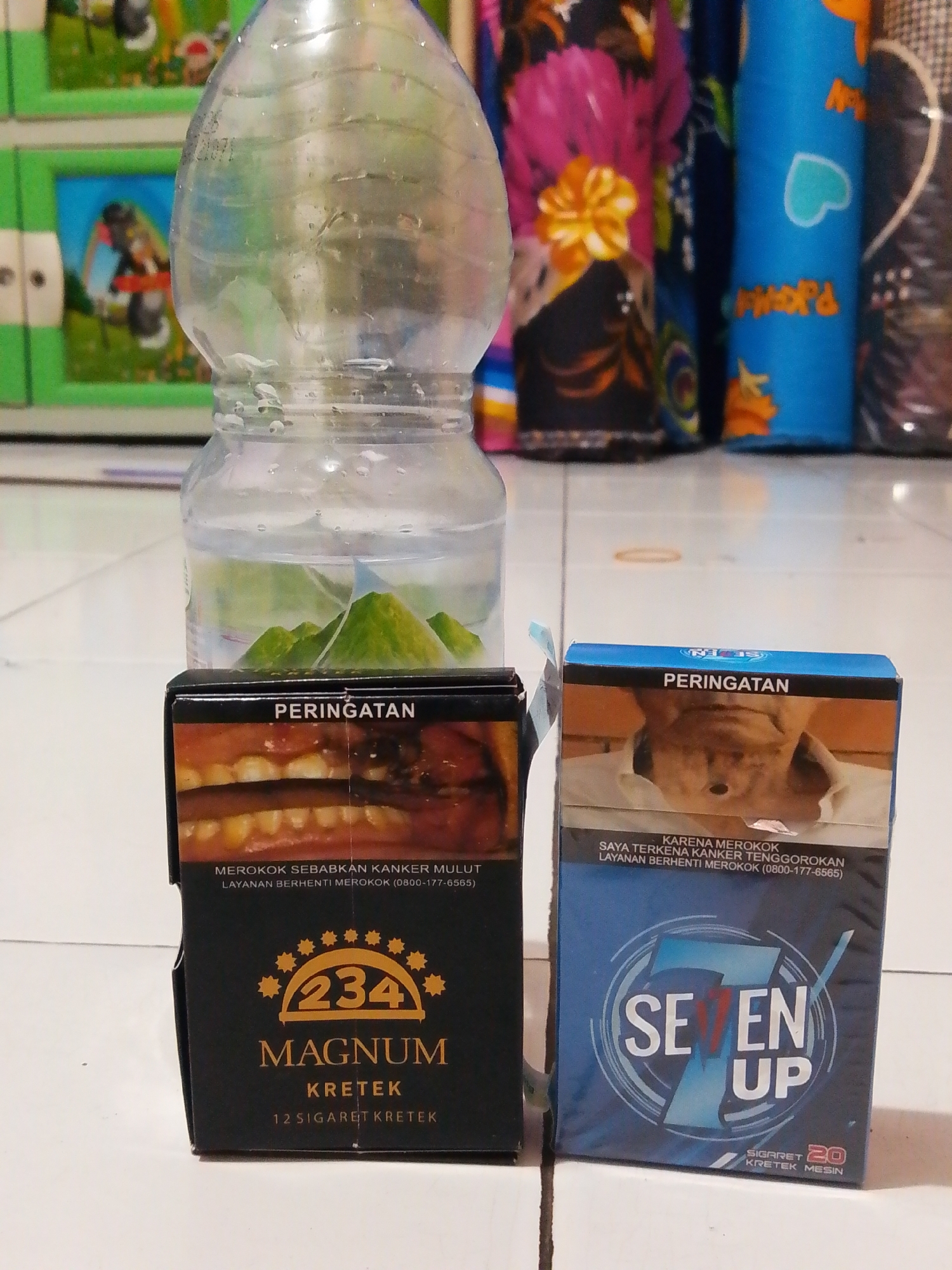 Two cigarette packs are placed on a tiled floor, in front of a plastic bottle containing water and some green mountainous scenery. The cigarette pack on the left is black and features an image of a mouth with rotten teeth, with the text "PERINGATAN" (Warning) at the top. Below the image, it reads "MEROKOK SEBABKAN KANKER MULUT" (Smoking causes mouth cancer) and "LAYANAN BERHENTI MEROKOK (0800-177-6565)" (Quit Smoking Service). The brand name "MAGNUM KRETEK" is displayed prominently with the number "234" in a circular design. It also states "12 SIGARET KRETEK" (12 Kretek Cigarettes).
The cigarette pack on the right is blue and shows a warning image of an elderly man's face, with "PERINGATAN" at the top. Below the image, it says "KARENA MEROKOK SAYA TERKENA KANKER TENGGOROKAN" (Because of smoking, I have throat cancer) and "LAYANAN BERHENTI MEROKOK (0800-177-6565)". The brand name "7 EN UP" is displayed with a stylized "7". It also indicates "SIGARET 20 KRETEK MESIN" (20 Machine Kretek Cigarettes). The background shows a blurred colorful pattern, likely decorative fabric or wallpaper. The scene is indoors, and the floor is made of white tiles.
