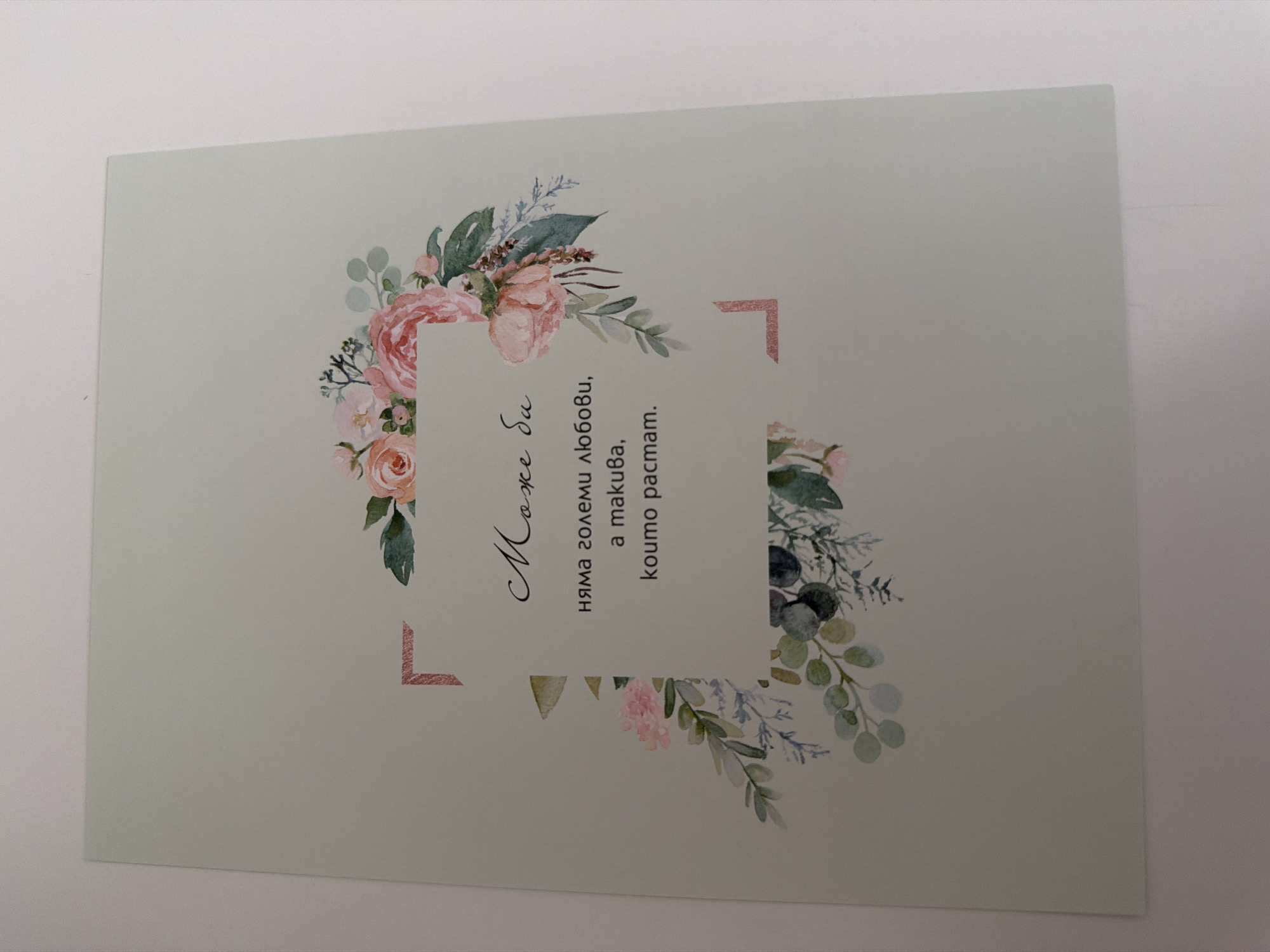 The image shows a light green card with a floral illustration. In the center of the card, there is text written in a script font and a more standard font. The script text reads "Може би", and the standard text reads "няма големи любови, a такива, koumo pactam.". The floral illustration features pink roses, eucalyptus leaves, and other greenery arranged in a bouquet. The card is placed against a white background. There is no visible connection to the city of Pazardzhik, Bulgaria in the image.