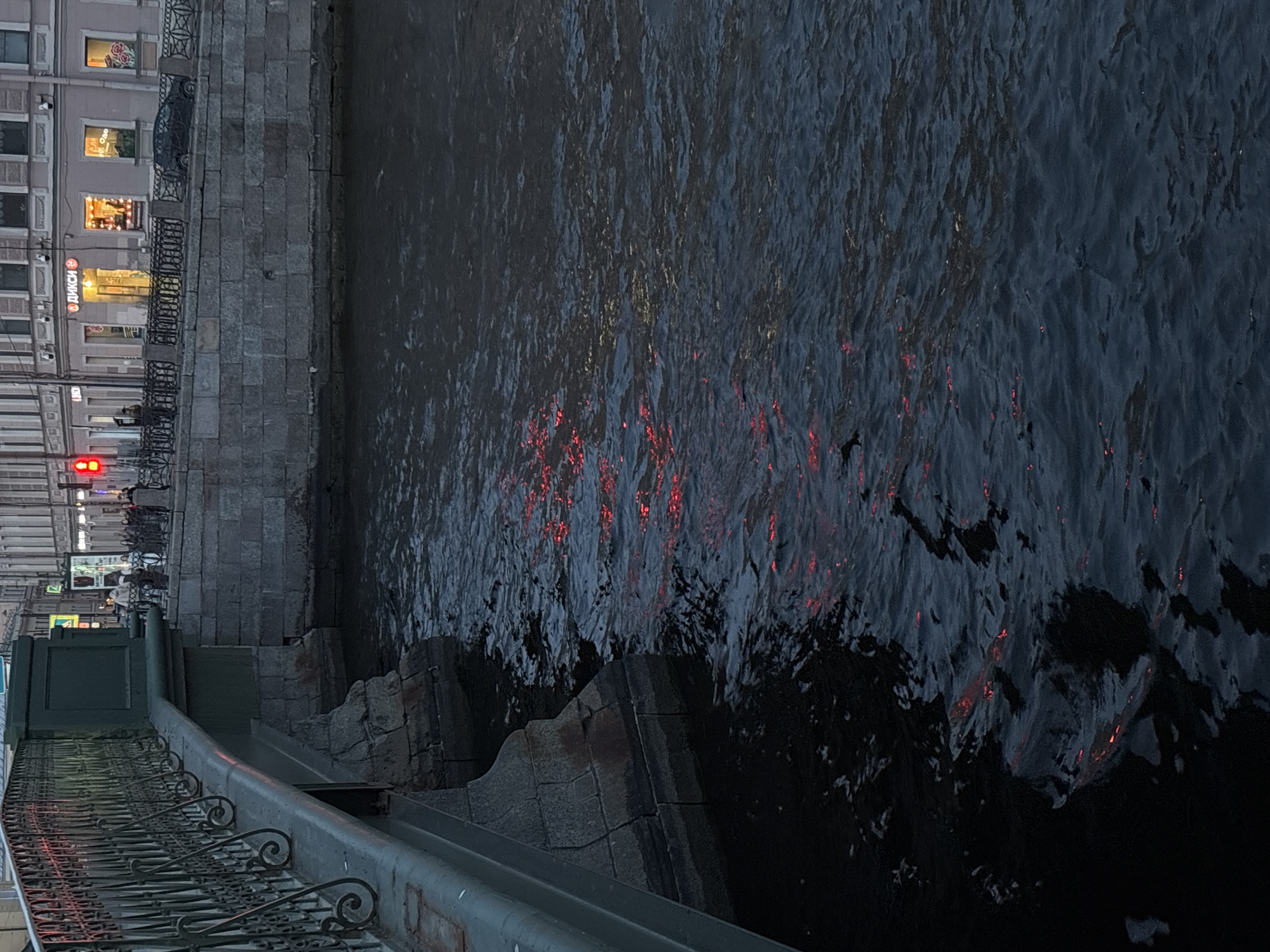 The image depicts a body of water, likely a canal or river, reflecting red light, possibly from nearby buildings or traffic lights. A stone embankment with steps leading down to the water is visible on the left. The embankment is partially obscured by a metal railing with decorative scrollwork. On the left side, buildings are discernible, suggesting an urban setting. A red traffic light is visible. The reflections on the water indicate some movement on the water's surface, causing ripples and distortions of the reflected light. The buildings on the left have lit windows, indicating it's likely evening. Given the presence of canals, stone embankments, and architecture, it is very probable that the location is Saint Petersburg, Russia. Specifically, it looks like the embankment of the Moyka River (Река Мойка). A sign "ДИКСИ" can also be seen on one of the buildings.