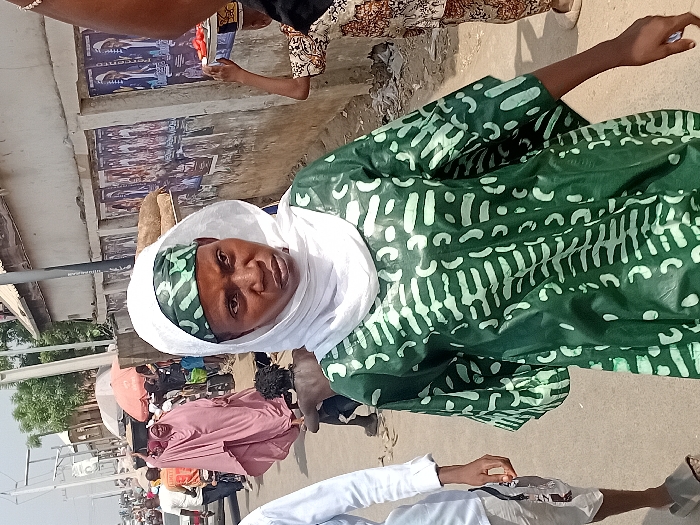 This image depicts a bustling street scene in Ogaminan, Nigeria. In the foreground, a dark-skinned woman with a direct gaze is positioned slightly off-center. She wears a loose-fitting green garment with light green patterned designs, a light green patterned headscarf, and an overlying white headscarf. Her left arm is visible, hanging by her side.
To the immediate left of the central figure, the lower torso and legs of an individual in a light blue long-sleeved shirt, light-colored shorts, and grey flip-flops are partially visible. Further back and to the left, a woman in a flowing pink or light purple robe is observed walking away from the viewer, appearing to carry an item. Behind her, a small, bare-chested individual with dark, curly hair is positioned closer to the ground. Additional indistinct figures and a vehicle are visible further down the paved street, suggesting ongoing movement and traffic.
On the right side of the frame, a light-colored concrete wall is visible, covered with multiple political campaign posters featuring male figures in suits and partially visible text, including "PERCEN". Several large, light-brown sacks are stacked against this wall. The arm and shoulder of a person are partially visible on the far right, holding a tray with sliced red produce (consistent with tomatoes) balanced on their head, indicating a street vendor. A dark, vertical street pole stands near the wall. The scene is illuminated by bright daylight.