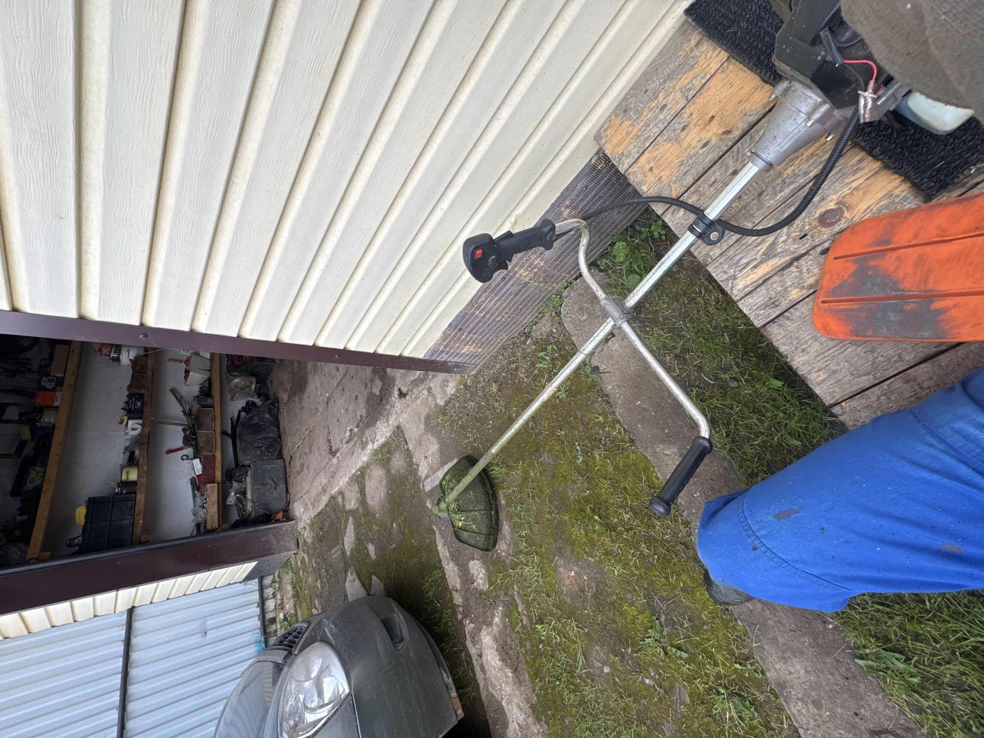 A first-person perspective shot shows a person wearing blue work pants, sitting on a wooden platform outside a garage or shed. In the foreground, to the left, is the person's leg and part of a grass trimmer, partially obscured by the person. The grass trimmer is silver and black, with a black handle and a red button. Further back, to the left of the trimmer, is the front of a dark gray car. The garage or shed has a light-colored, horizontal siding and an open doorway revealing shelves stocked with various items, including containers, tools, and bags. The ground is a mix of concrete, dirt, and patchy green grass, with some puddles indicating recent moisture. The overall scene suggests a residential or utility area, possibly in Borodulino, Russia, where yard work is either about to commence or has recently been completed.