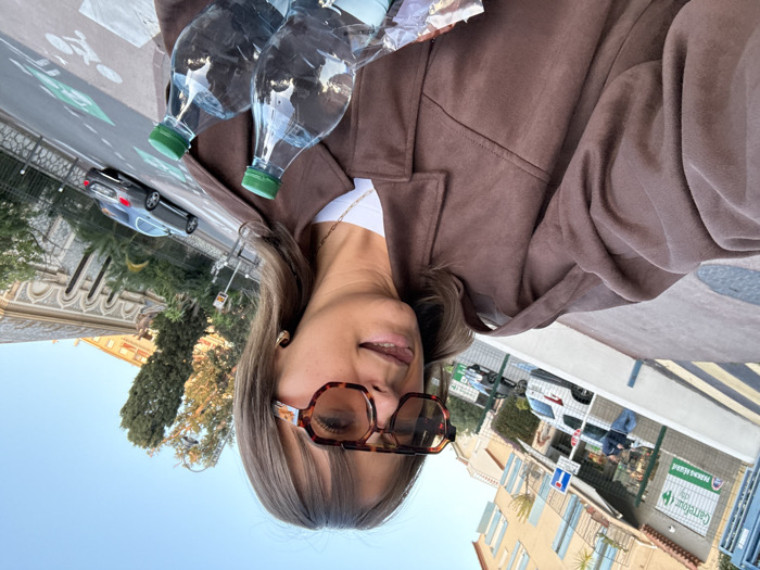 A woman wearing tortoise shell sunglasses and a brown jacket poses for a photo, holding two water bottles. A street scene is visible in the background with buildings, palm trees, a parked car, and a bicycle lane. A Carrefour store sign is also visible. The sky is a light blue. The woman's hair is brown with blonde highlights. She wears a gold necklace and small hoop earrings.