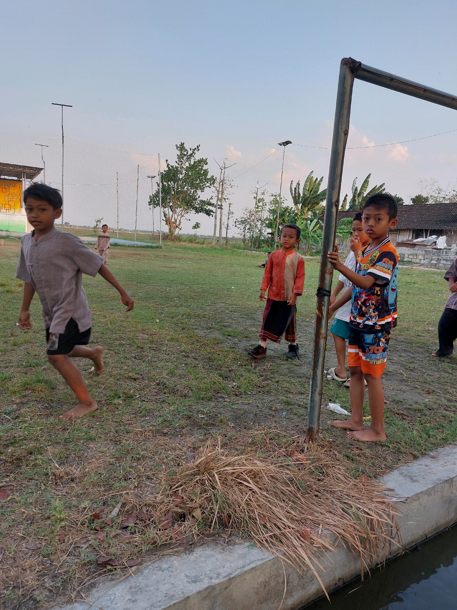 The image shows an outdoor scene in what appears to be a rural area or a sports field, with a blue sky and sparse vegetation. Several young boys are present. In the foreground, a boy with dark hair is running to the left, barefoot, wearing a light brown shirt and dark shorts. To his right, a boy with dark hair is standing near a metal pole, wearing a colorful t-shirt with an orange and black design and matching shorts. He is holding onto the pole. Behind him, another boy is partially visible, wearing a white t-shirt and green shorts. Further back, to the left, a boy in an orange and brown outfit with a dark skirt-like garment is standing. In the background, there is a structure with a white and yellow facade, netting that extends upwards, trees, and some buildings with tiled roofs. A concrete edge and a body of water are visible in the lower right corner. The ground is covered with dry grass and some leaves.