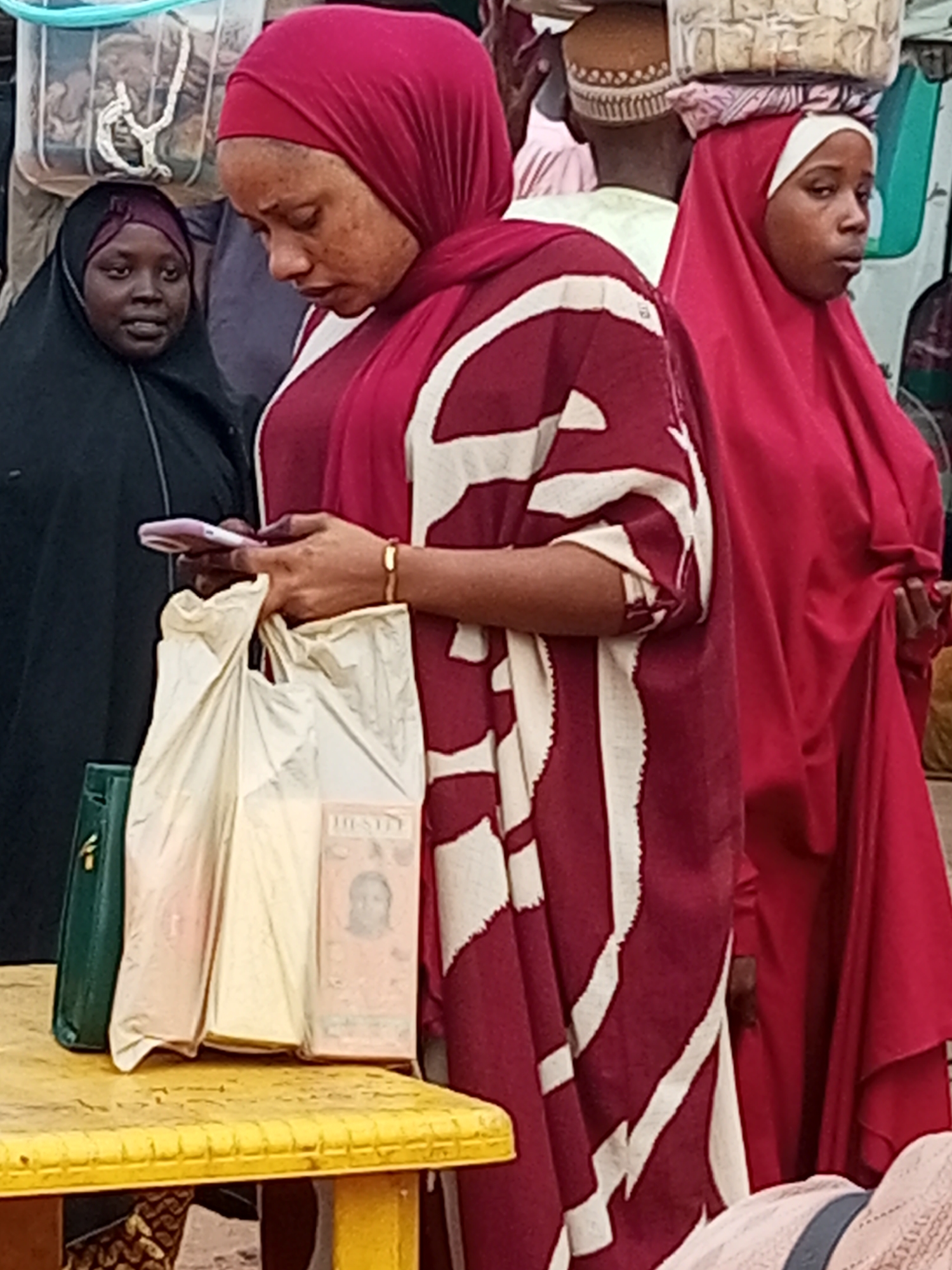 In Gombe, Nigeria, an outdoor market scene unfolds. A woman in a red hijab and a maroon and white patterned garment is engrossed in her smartphone, holding a light pink phone and a light beige shopping bag. Next to her, another woman in a black hijab is visible from the shoulders up. To the right, a third woman in a red hijab and garment stands facing away. A yellow table is in the foreground, laden with a green bag, the beige shopping bag containing a small booklet with an image of a person, and other indistinct items. In the background, a man wearing a woven hat and a light green shirt is partially visible. The atmosphere suggests a bustling market environment.