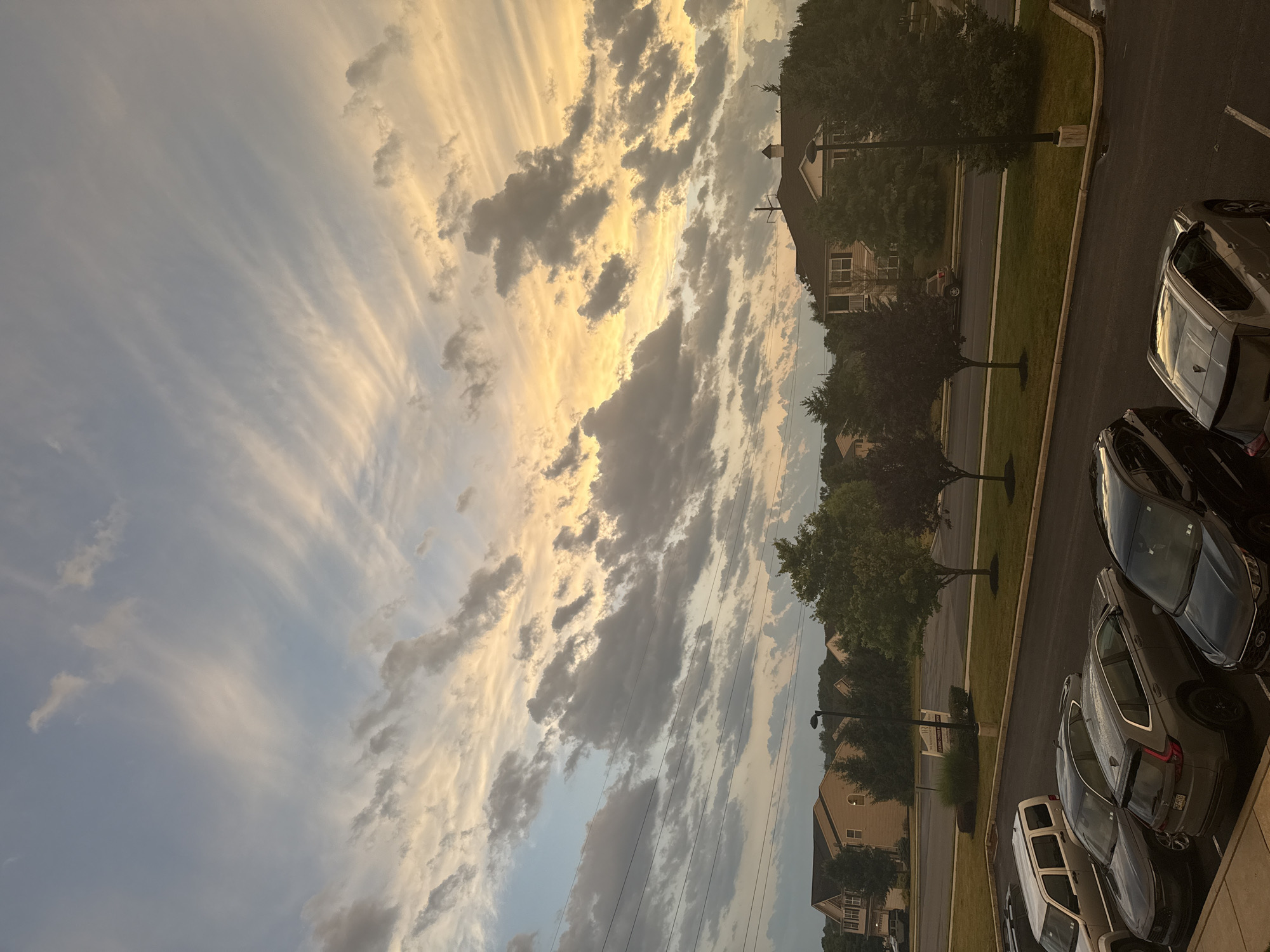 This is an exterior, eye-level, vertical shot of a suburban street and parking lot at dusk, in Allen Township, United States of America. The sky is filled with dramatic, wispy clouds illuminated by the setting sun in hues of yellow and orange, contrasted with patches of blue. Below the sky, a street lined with trees and houses can be seen. Several cars are parked in a lot in the foreground, including a silver SUV, two dark-colored SUVs, and a black sedan. The parking lot surface is asphalt, and there are yellow parking lines. The houses are two-story structures with beige siding. A lamppost stands near the street. The overall scene suggests a quiet evening in a residential area.