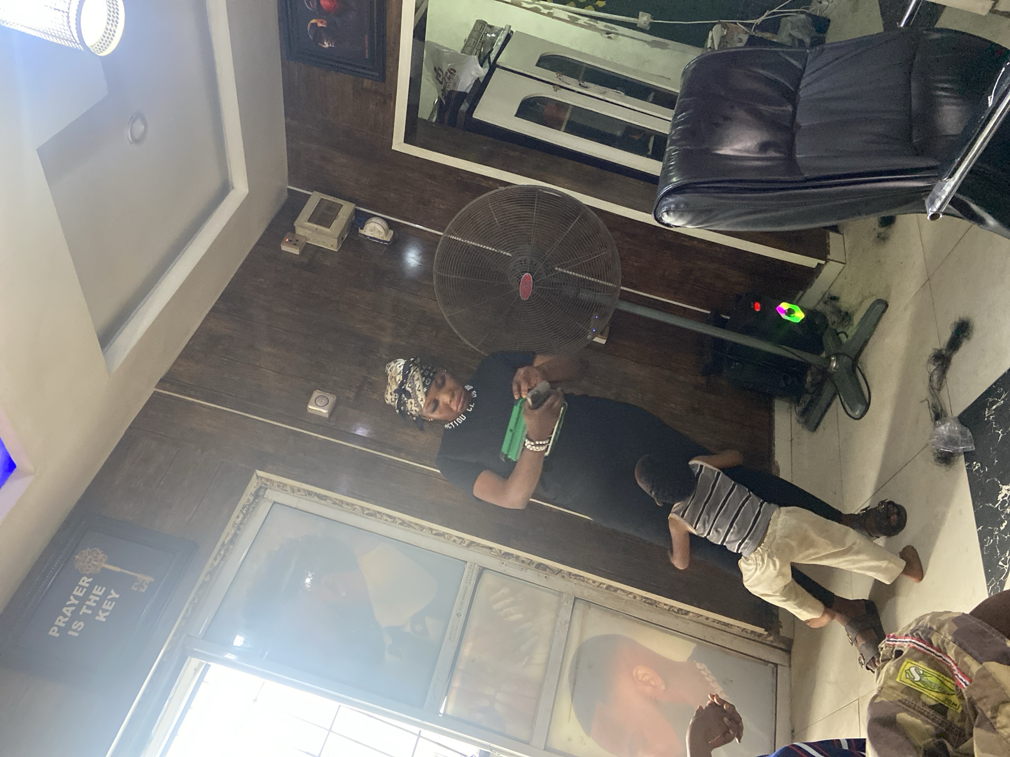 The image captures an indoor scene, likely a waiting area or a salon, during the daytime, suggested by the bright light filtering through a window on the left. The location context indicates it's in Lokoja, Nigeria.
In the center, an adult woman, wearing a black t-shirt and a patterned head wrap, stands holding a bright green object, possibly a book or phone, in her hands and looking down at it. Text on her shirt is partially visible, appearing to say "LECTION LL". Beside her, a small child, likely a toddler, wearing a striped tank top and light-colored trousers, stands facing her, seemingly observing what she is doing. Part of another person, clad in a patterned top, is visible seated in the bottom right corner, suggesting more people are present in the room.
The room features dark wooden paneling on the walls, providing a warm aesthetic. A large black standing fan is positioned behind the woman, indicating a need for air circulation. To the right, a black leather salon or office chair is visible. The floor is a light, possibly tiled or laminated, material.
Notable decorative elements include a framed picture on the left wall featuring a golden key and the text "PRAYER IS THE KEY". Another framed picture is visible higher up on the wall, partially obscured. A modern-style ceiling light fixture hangs from the ceiling. A mirror reflecting a door is mounted on the far wall. Electrical outlets and switches are also visible on the wooden wall paneling.
