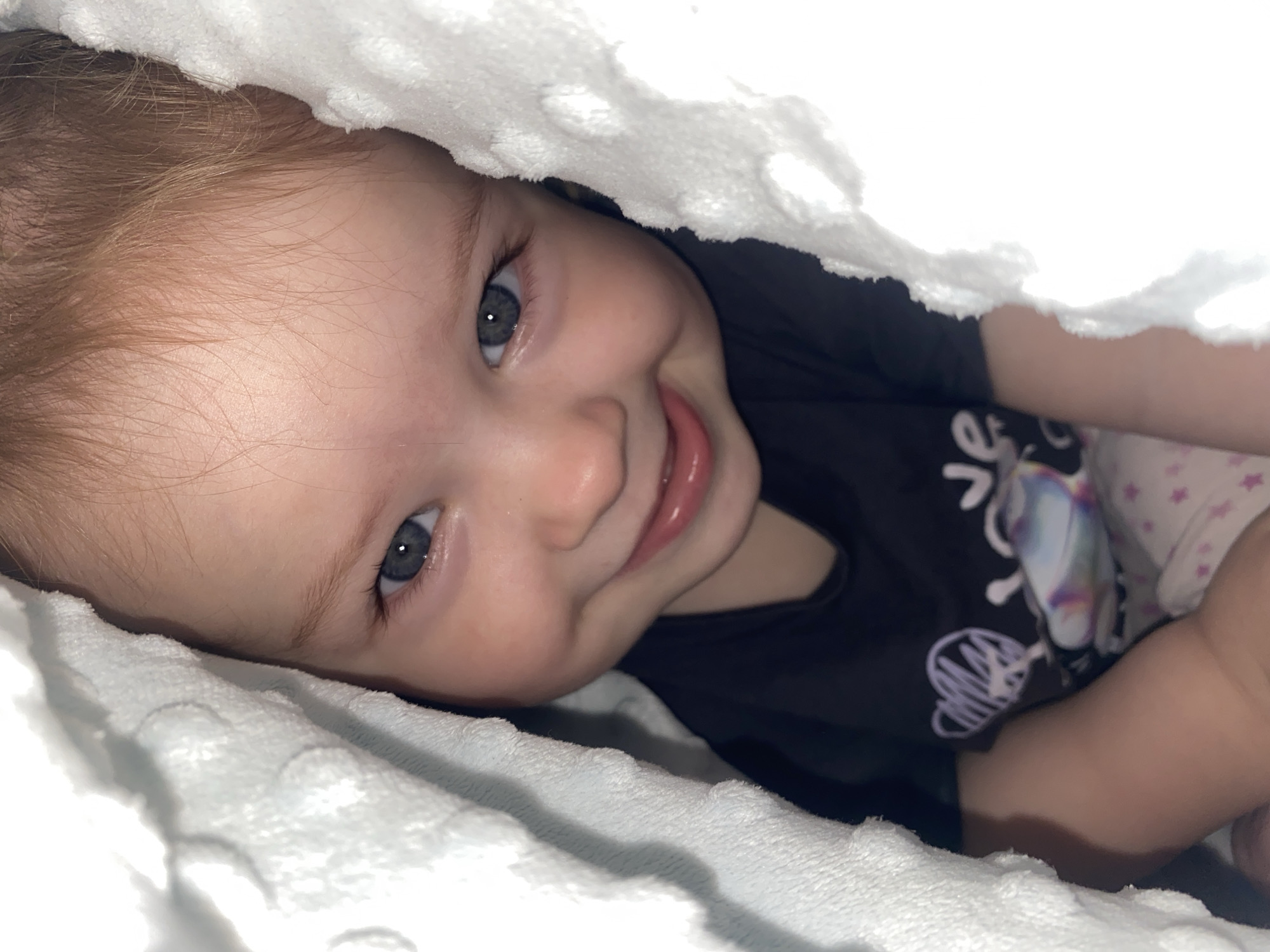 A close-up, high-angle shot captures a smiling baby or young child peeking out from under a soft, white textured blanket. The child's head is tilted slightly, and they look directly at the viewer with wide, clear blue-gray eyes. They have fair skin, light reddish-brown hair visible around their forehead, and a bright, open-mouthed smile, showing part of their gums and possibly a few emerging teeth.
The child is wearing a dark-colored (likely black) t-shirt, on which white graphic designs and letters are visible. One design appears to be a stylized heart or abstract shape, and some letters, possibly "Je" or "Love," are partially seen. A section of another garment, light-colored with pink star patterns, is visible near the child's right arm.
The white blanket, possibly made of minky fabric due to its textured, dotted surface, frames the child's face and extends into the foreground and background, creating a cozy and intimate atmosphere. The lighting is soft, highlighting the child's features and the blanket's texture. The background behind the child's body is dark and indistinct, suggesting a dimly lit space or the shadow cast by the blanket. The overall mood is one of joy and playfulness.