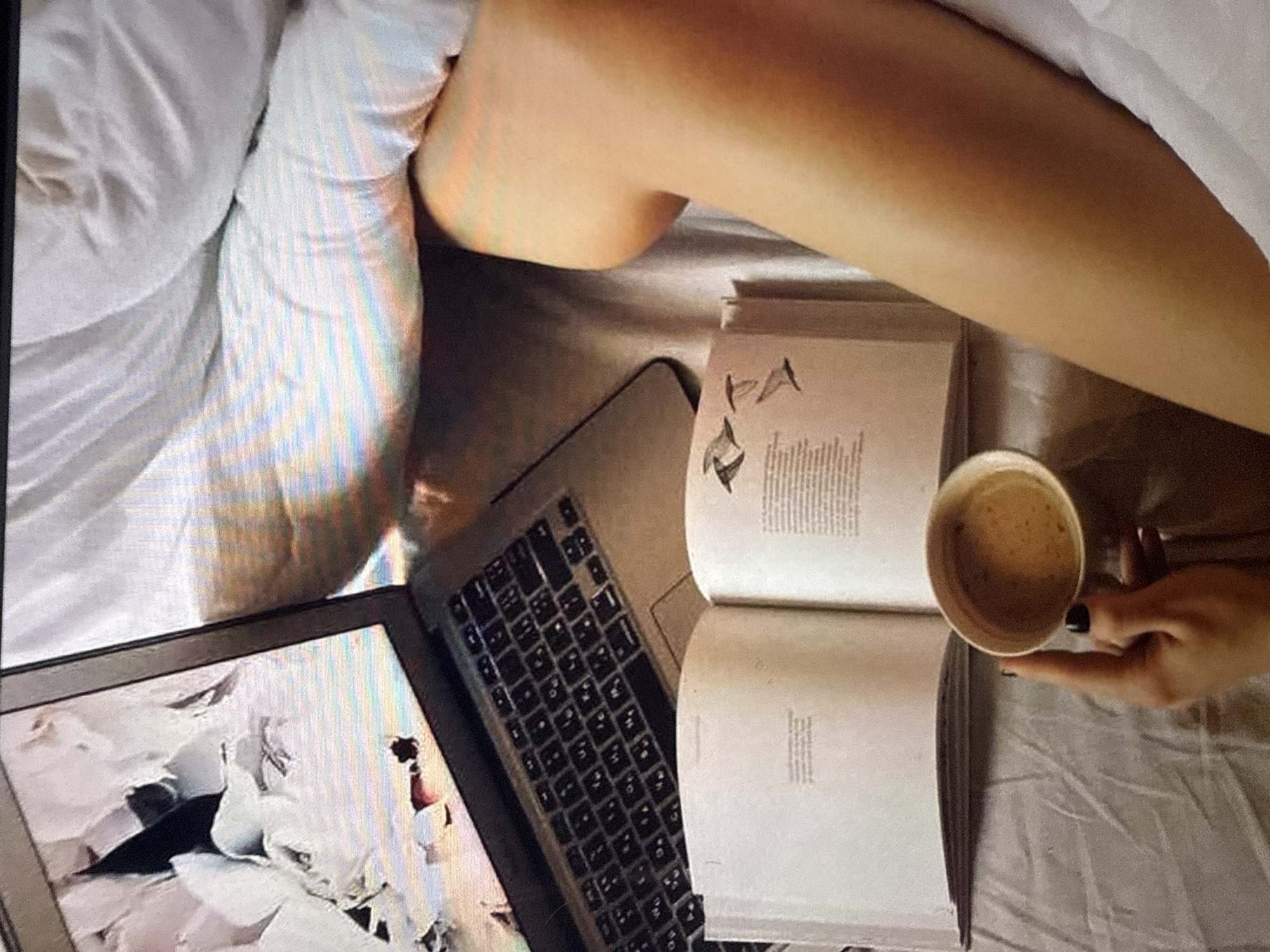 An over-the-shoulder, eye-level shot shows a person holding a white coffee cup with a light brown beverage and a finger with black nail polish. The coffee cup is positioned above an open book with white pages. The book is lying on a surface that appears to be a bed covered with white linens. To the left of the book, a silver laptop is visible, open and displaying an abstract image on its screen. The laptop is positioned on the white linens. The background consists of white bedding, creating a soft, cozy atmosphere. The lighting is natural, possibly from a window, and casts subtle shadows. The image gives a sense of a relaxed, private moment, possibly a morning or evening routine.