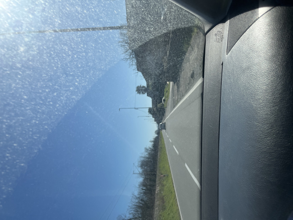 The image depicts a daytime scene from the interior of a vehicle, looking forward through a windshield. The windshield exhibits numerous dirt specks and streaks, particularly concentrated in the upper right quadrant, with sunlight causing significant glare.
The immediate foreground shows a dark, textured dashboard. In the distance, a paved two-lane road extends into the frame. The road features a solid white line on its right edge and a dashed white line separating the two directions of travel. A light-colored, possibly white or silver, small car is visible in the far lane, moving away from the viewer.
To the left of the road, a green grassy embankment is present, bordered by a dense line of bare trees or bushes. Overhead, utility wires or power lines are visible running parallel to the road. A small, reddish-orange marker pole is situated on the embankment.
To the right of the road, a rough, unpaved shoulder transitions into an area with what appears to be a low, dark-colored stone or concrete wall or structure, partially obscured by leafless vegetation. Several tall streetlights with black poles are spaced along this side of the road, receding into the distance. The sky is clear and bright blue, indicating sunny weather. No people are discernible in the scene. The location is Bistrica, Montenegro.