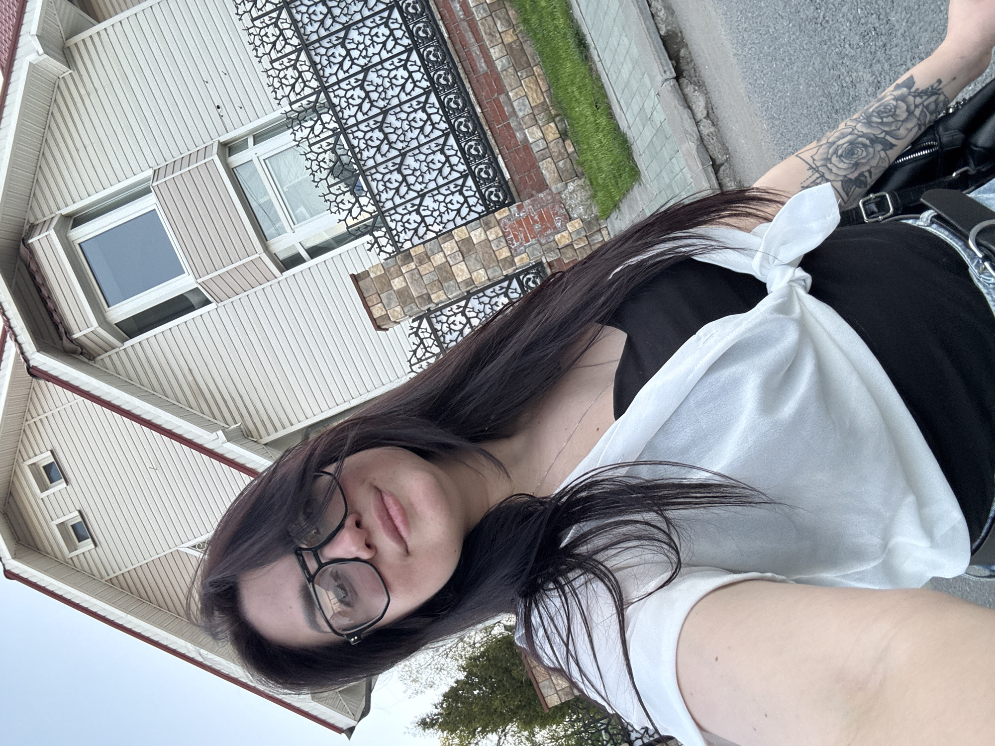 A woman with dark hair and glasses is taking a selfie outside in front of a house. She is wearing a black top with a white shirt tied around her waist. Her left arm is visible and features a tattoo. Behind her is a house with light-colored siding and trim, and decorative metalwork adorns part of the facade. There is also a sidewalk and some greenery visible in the background.