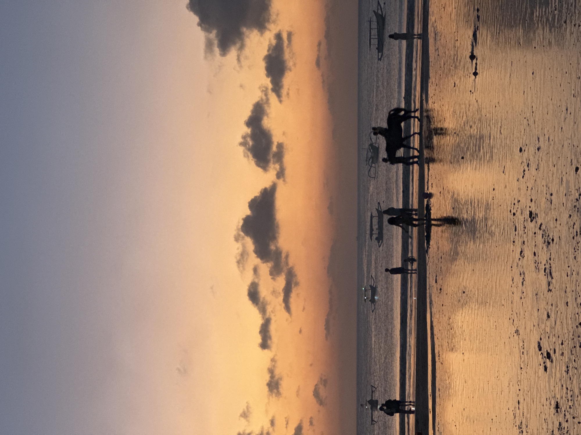 The image depicts a serene beach scene in Kuta Selatan, Indonesia, at dusk. The sky transitions from a soft lavender at the top to a warm orange hue near the horizon, with scattered, dark clouds. The sea is calm, with several traditional Indonesian boats (jukung) visible in the distance. On the wet sand of the beach, the reflections of the sky and boats create a shimmering, golden surface.
In the foreground, silhouettes of people are scattered across the beach. A notable group includes a person riding a horse, with another person standing next to the horse, perhaps preparing to mount or dismount. Further down the beach, other individuals and smaller groups are either walking or standing, their figures indistinct against the twilight glow. The wet sand, speckled with small debris, catches the fading light, creating a reflective, mirror-like effect. The overall atmosphere is peaceful and tranquil, characteristic of a tropical sunset.
