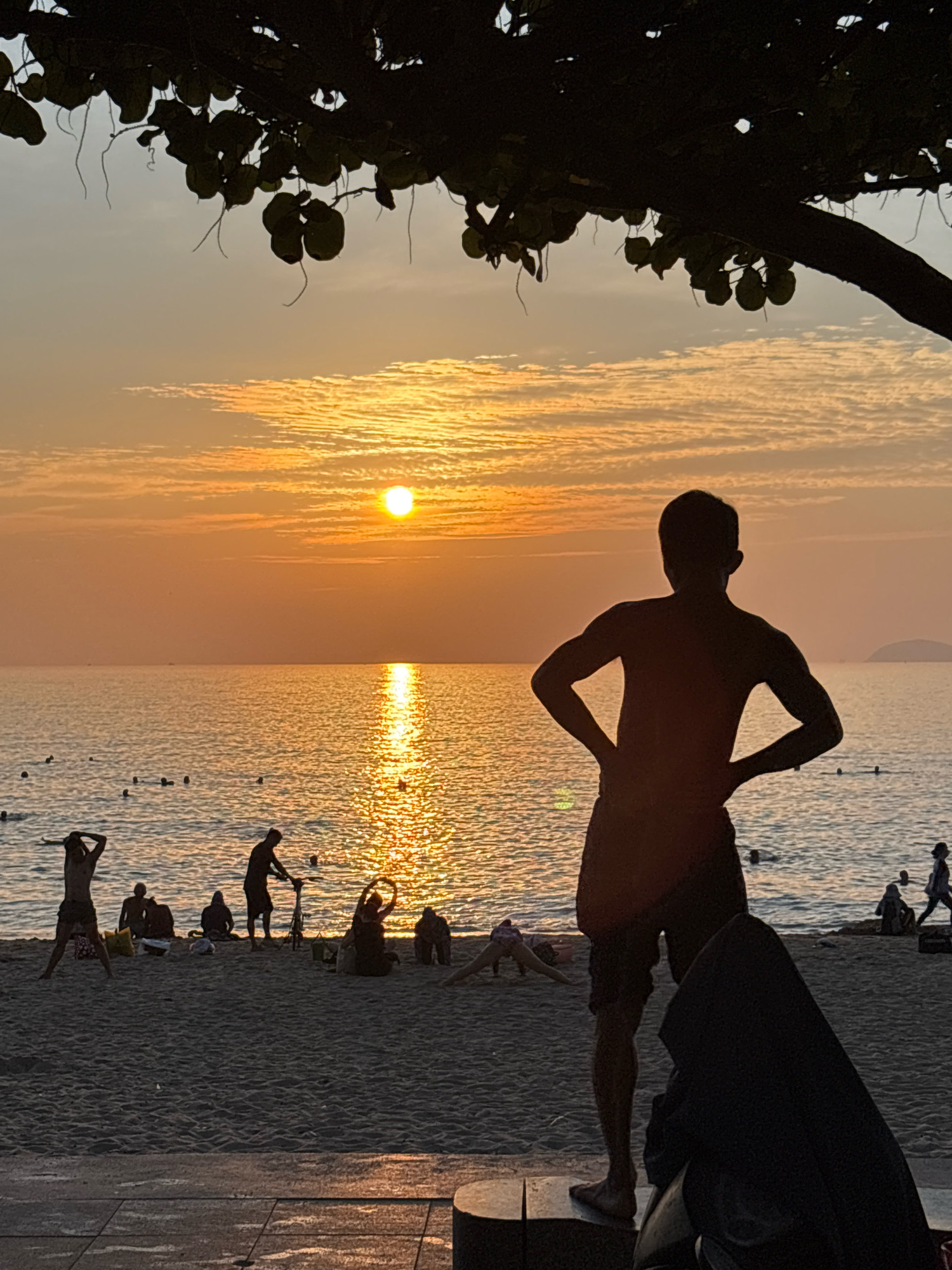 The image depicts a serene sunset scene at a beach in Nha Trang, Vietnam. The sun, a bright orange orb, is low on the horizon, casting a golden reflection across the calm sea. The sky is adorned with scattered clouds, painted in shades of orange and yellow.
In the foreground, silhouetted against the vibrant sunset, stands a young man with his hands on his hips, his back to the camera. He is wearing dark shorts and appears to be enjoying the view.
Further down the beach, other individuals are visible in silhouette as well. Some are standing, others are seated or lying down, suggesting a relaxed atmosphere with people enjoying the end of the day. A few individuals are in the shallow water, with some appearing to be swimming or wading.
A bicycle is also visible on the sand, near a group of people. The beach itself is sandy, with some areas appearing to be paved or have a walkway in the immediate foreground. The overall impression is one of a peaceful evening at a popular tourist destination. The presence of the sea, the beach, and the silhouettes of people enjoying leisure activities strongly suggests a location within Nha Trang, known for its beautiful coastline and vibrant beach life.