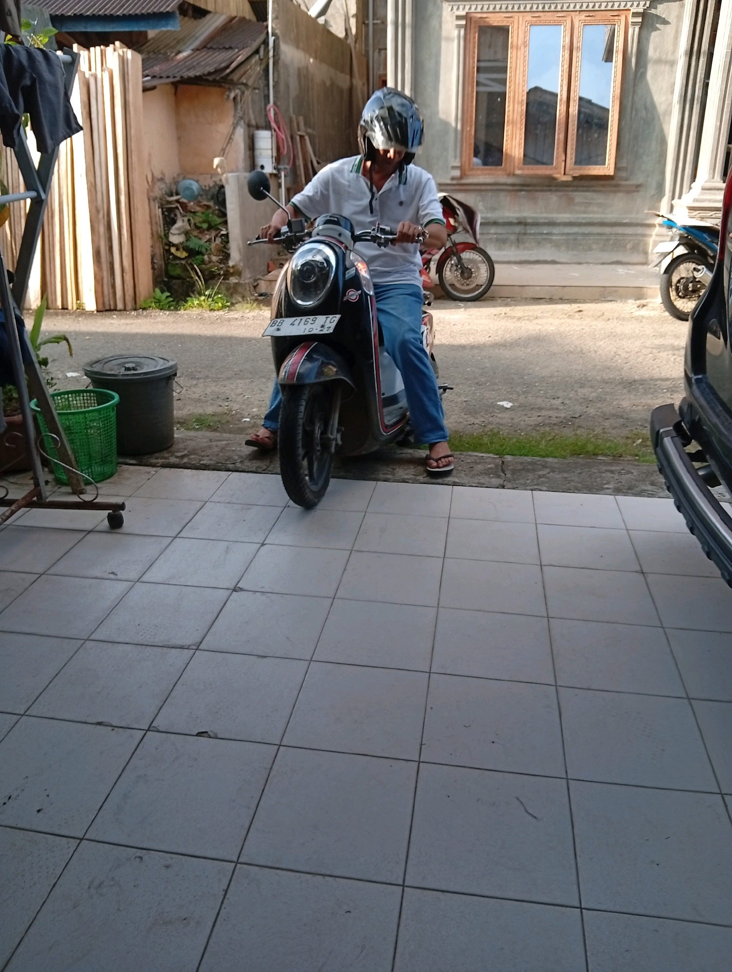 A person wearing a helmet and blue jeans sits on a scooter in front of a building. The scooter has a license plate that reads "BB 4169 IG". The building has a wooden window with three panels and a grey facade. In the background, there are stacked wooden planks and a green basket next to a black trash can. The scene appears to be in a residential area within the City of Gunungsitoli, Indonesia, possibly on a paved street or driveway, with a tiled floor in the foreground.