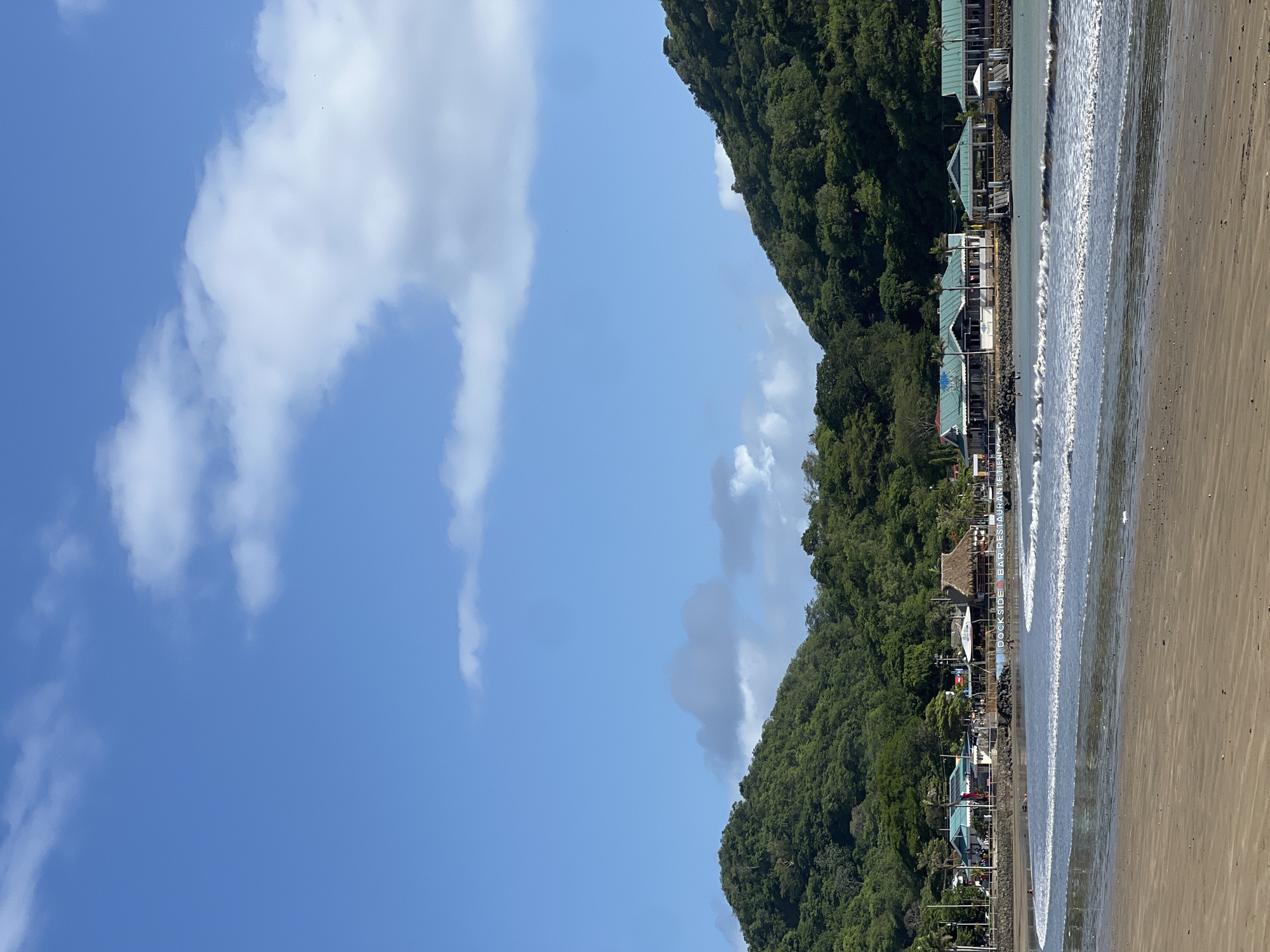 This is a photograph taken at the San Juan del Sur beach in Nicaragua, looking west towards the ocean from the shore. The foreground shows a wet sand beach with gentle waves breaking. In the mid-ground, there is a row of buildings situated along the coastline, featuring a mix of architectural styles including thatched roofs and modern designs with blue roofs. These appear to be restaurants or businesses. A seawall is visible between the buildings and the water. Some people are present on the beach and in the water, engaged in activities like swimming or lounging. The background is dominated by lush green, tree-covered hills on either side of the bay, with a bright blue sky with scattered white clouds above. The overall scene depicts a sunny day at a coastal town.
