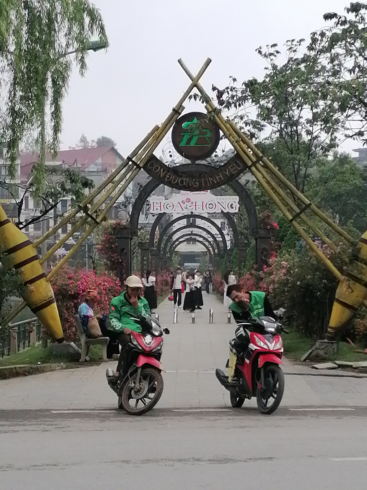The image captures a scene in Sa Pa Ward, Vietnam, featuring two men on red motorcycles at the entrance of an archway. The archway is decorated with bamboo structures, greenery, and signs written in a local language, possibly Vietnamese, that include the phrases "CON DUONG TINH YEU" and "HOA HONG". Beyond the archway, a pathway is lined with more arches, flowers, and people strolling along. One man on a motorcycle is wearing a green vest and a white cap, holding a hand to his face. The other man on a motorcycle also wears a green vest and is looking to his right. In the background, there are buildings and trees, with an overall foggy or overcast sky.