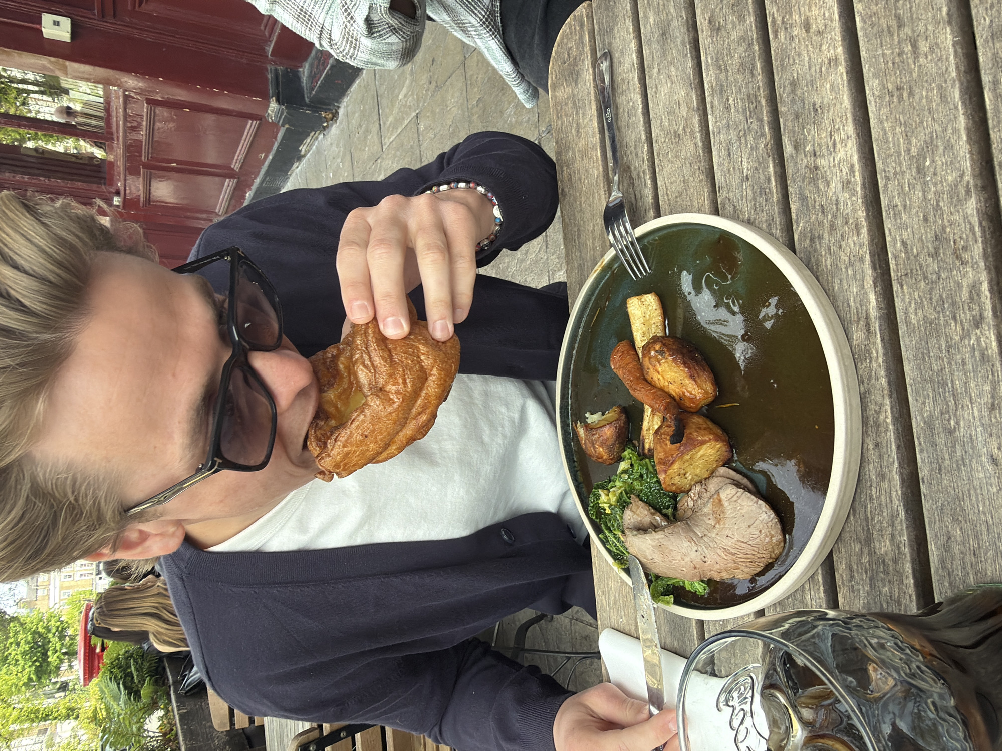The image shows a person eating a meal at an outdoor table in what appears to be London, United Kingdom. The individual, who seems to be a young man, is fair-skinned, wearing sunglasses, a white t-shirt, and a dark blue cardigan. He is in the process of taking a large bite out of what looks like a Yorkshire pudding. On the table, there is a plate containing what appears to be a Sunday roast with meat, potatoes, parsnips, and green vegetables, likely kale. A fork is resting on the plate and there is a knife visible by the plate. A clear glass is partially visible at the bottom right of the frame. The table itself is made of wooden planks. Behind the person, a glimpse of the city landscape is seen, including a red phone booth and foliage, and a brick building. The scene is likely a restaurant or pub with outdoor seating.