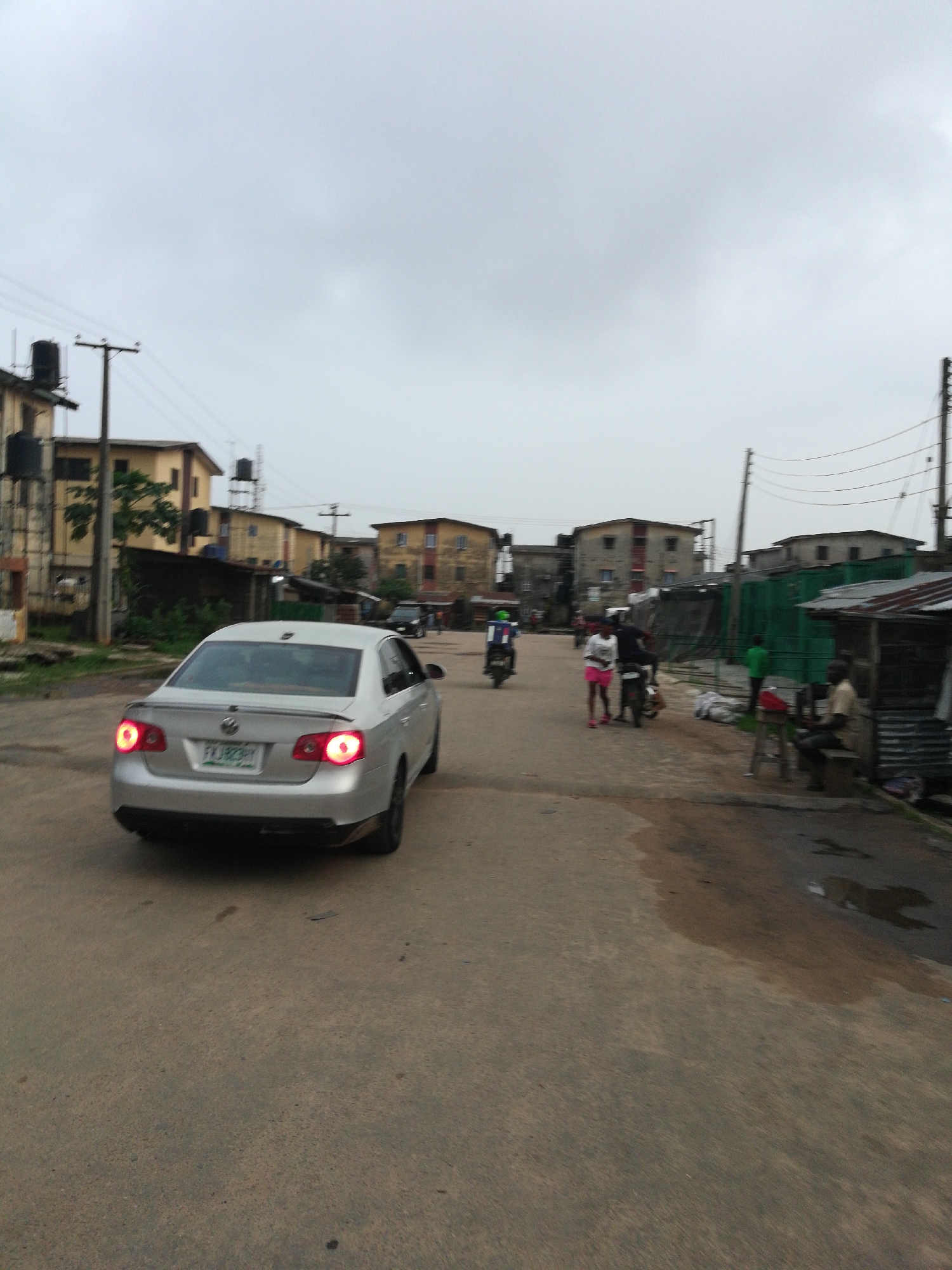 The image depicts a street scene in Unknown, Unknown under a cloudy sky. In the foreground, a silver car is driving away from the viewer. In the background, a person is riding a scooter on the road. Several pedestrians are on the side of the road, near makeshift structures. There are various buildings visible in the background, with water tanks on top. Overhead power lines are strung between utility poles. The road surface is weathered and shows signs of wear.