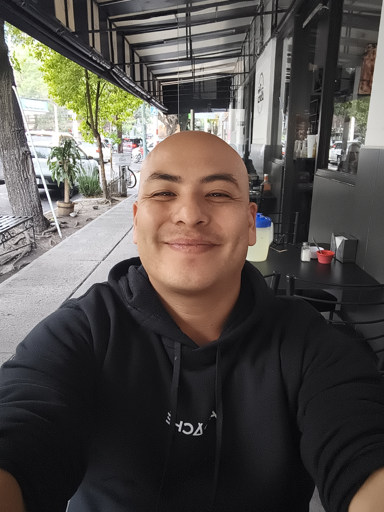 A selfie of a bald man wearing a black hoodie outdoors in Mexico City. He is seated at a table outside a restaurant or cafe, with trees, street infrastructure, and cars visible on the street. The cafe has a black awning and black-framed windows.