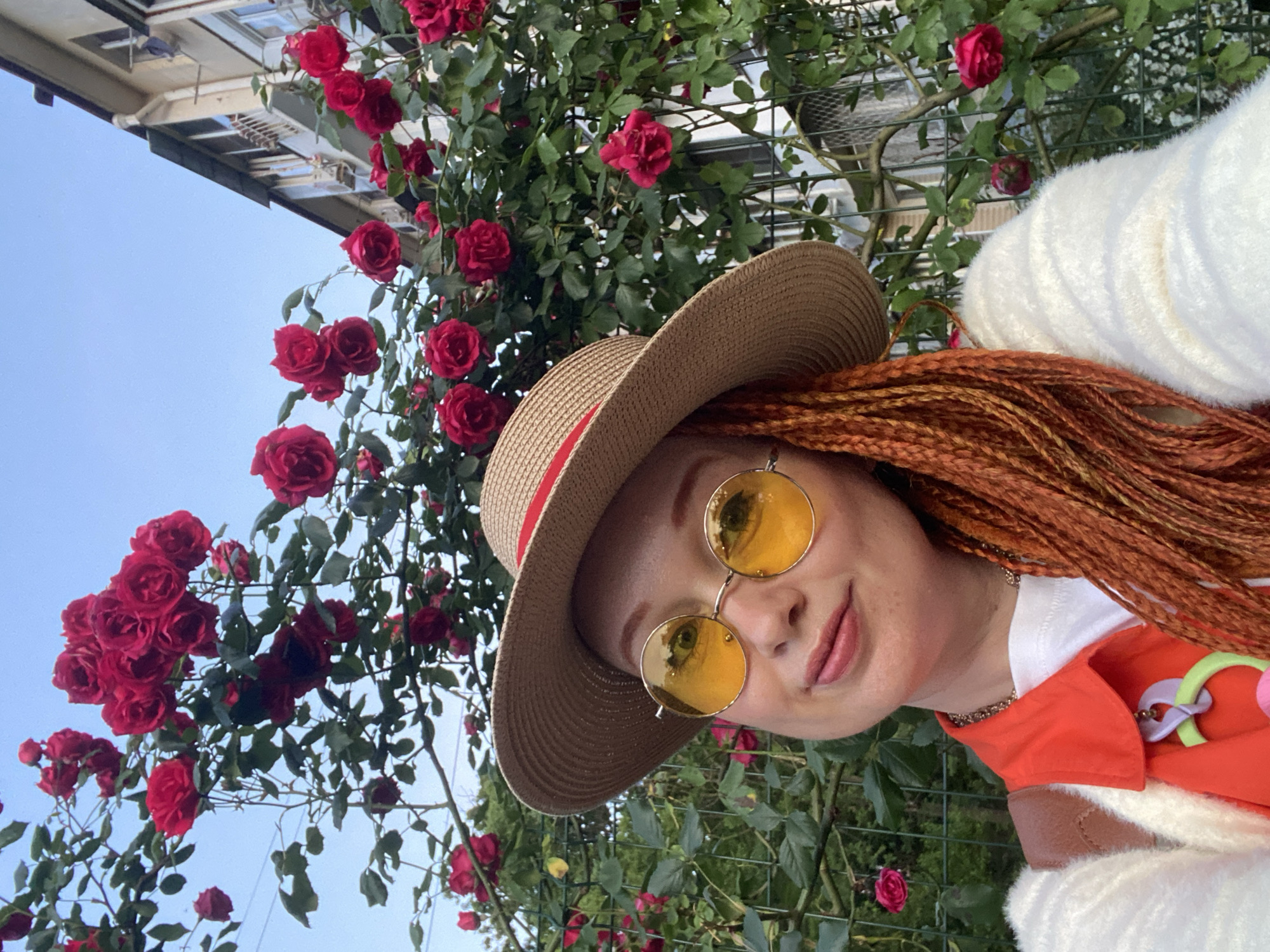 This selfie captures a woman posing in front of a vibrant display of red climbing roses under a clear blue sky, likely in Sochi, Russia. The woman, positioned in the foreground, has long, reddish-brown braided hair styled into dreadlocks or box braids. She wears a straw hat with a red band, and large, round sunglasses with yellow-tinted lenses, which reflect her eyes. A gentle, pleasant smile plays on her lips.
She is dressed in a white top, an orange outer garment (possibly a jacket or vest) featuring a unique, colorful accessory with purple, green, and pink elements, and a fluffy white garment draped over her right shoulder. A delicate gold chain necklace is visible around her neck.
The background is dominated by lush rose bushes laden with numerous clusters of deep red roses and their green leaves, supported by a metallic trellis. In the upper left portion of the image, a light-colored building or balcony structure is partially visible against the bright, unclouded sky. The bright daylight suggests either morning or afternoon on a clear, sunny day. The overall mood is cheerful and serene, conveying a sense of enjoyment in a beautiful garden or outdoor setting. No text is visible in the image.