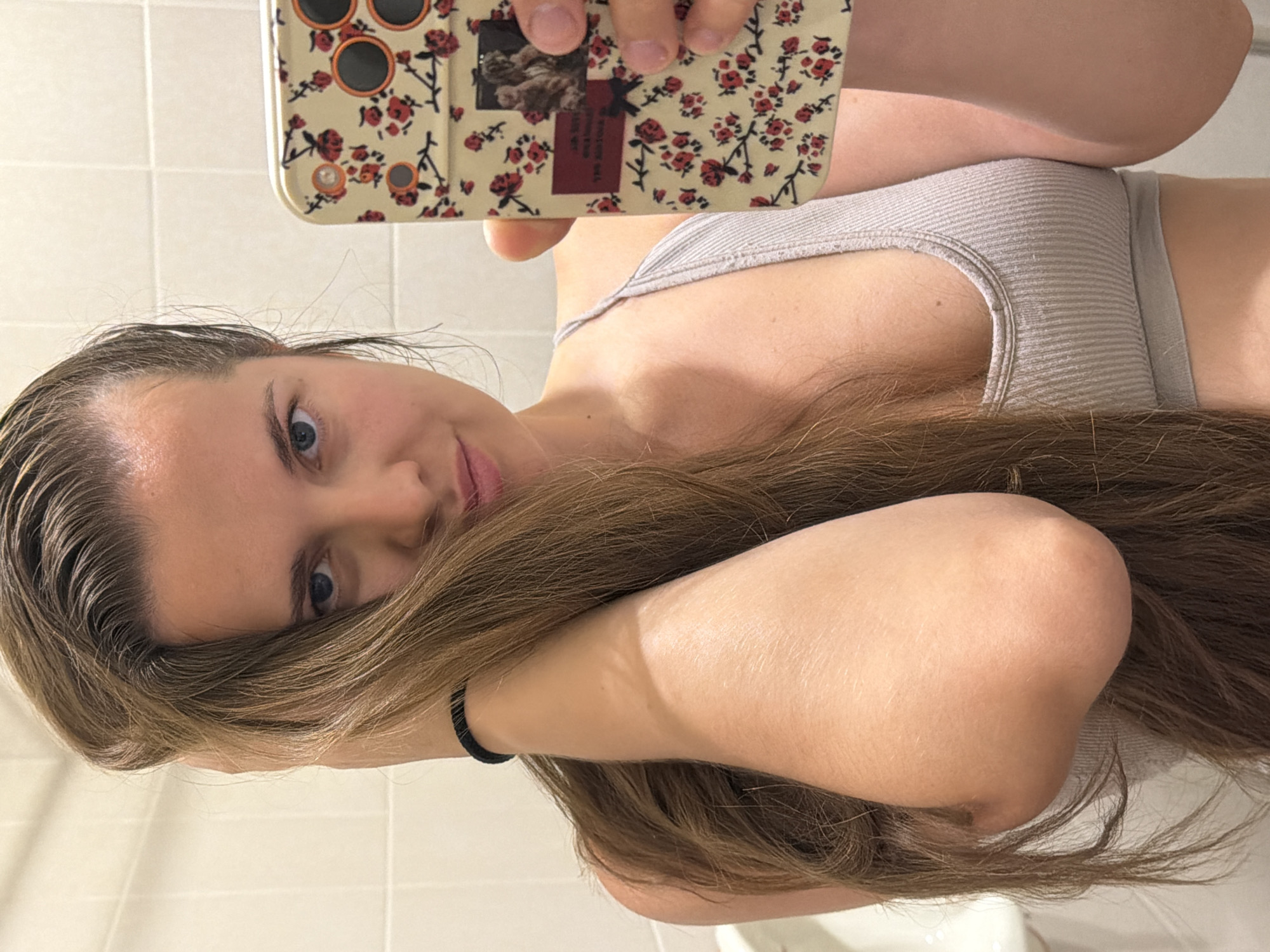 A woman is taking a selfie in what appears to be a bathroom. She is holding her phone in her right hand, and her left arm is bent, with her hand resting on her head and hair. She is wearing a ribbed, light-colored sports bra. The phone case has a floral pattern with red roses and green leaves. The background consists of white tiled walls. The overall lighting suggests an indoor setting. The image does not contain any information that would allow for the identification of a specific location within the city of Sochi, Russia.