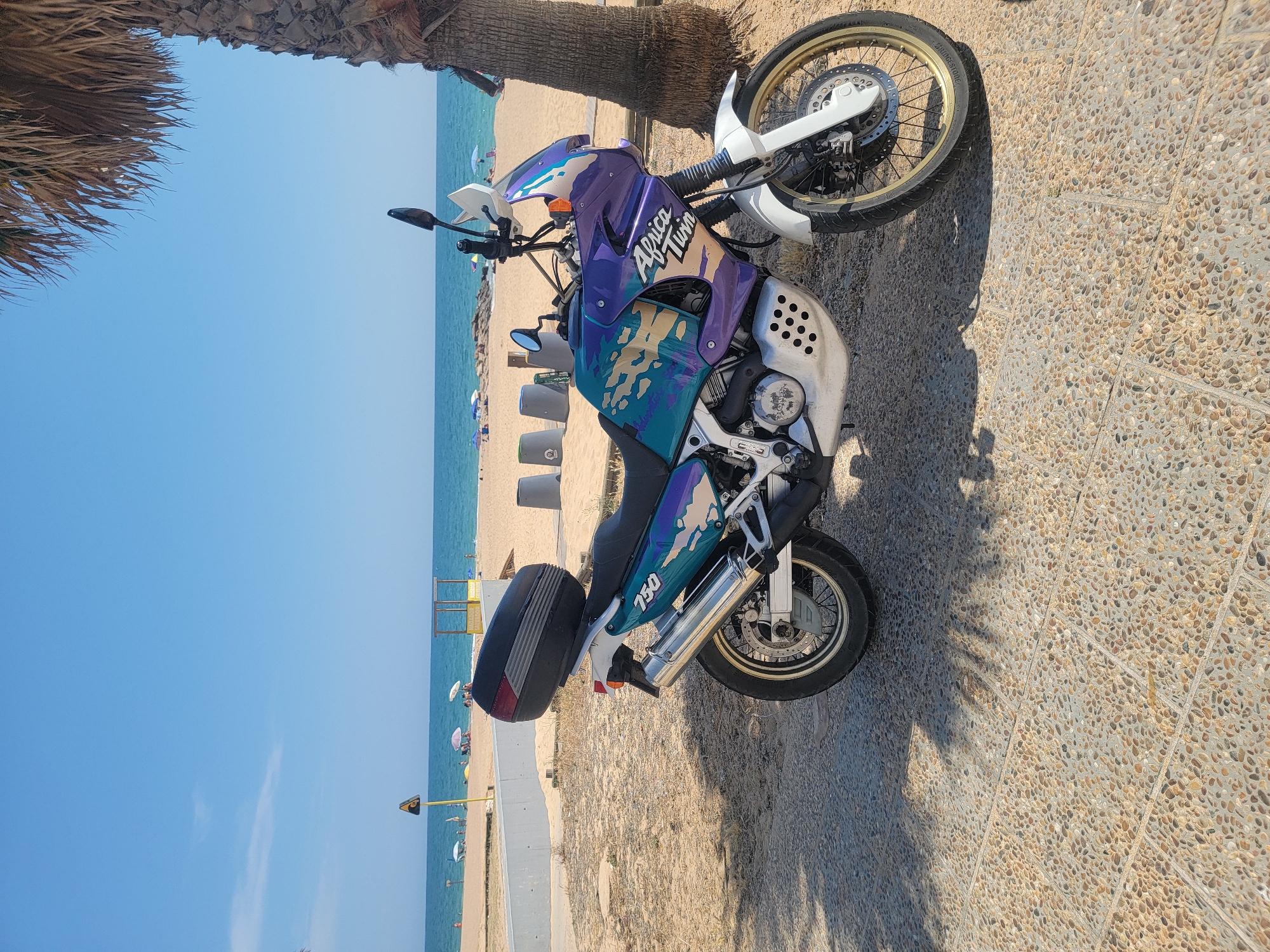 A Honda Africa Twin motorcycle, with a distinctive purple, teal, and gold livery, is parked on a paved walkway. The motorcycle is equipped with a black top case and a silver exhaust system. In the background, a sandy beach with scattered beach umbrellas and people is visible, leading to the blue ocean under a clear sky. A palm tree trunk stands to the right of the motorcycle, casting a shadow on the walkway. The scene appears to be a coastal area in Quarteira, Portugal.