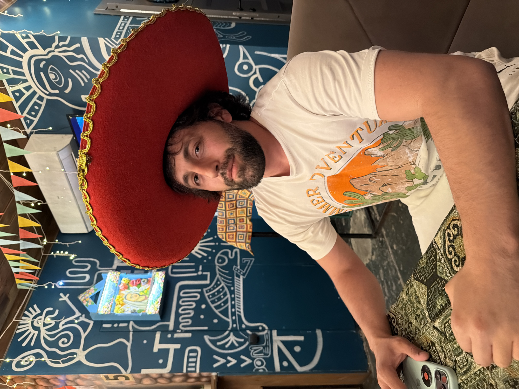 A person with a beard and a large red sombrero sits indoors, holding a smartphone in their right hand. They are wearing a white t-shirt with a colorful graphic. The background is a blue wall decorated with white drawings and patterns, along with colorful hanging decorations. The subject appears to be sitting in a restaurant, potentially in Krasnoyarsk, Russia, based on the presence of typical local decorations.