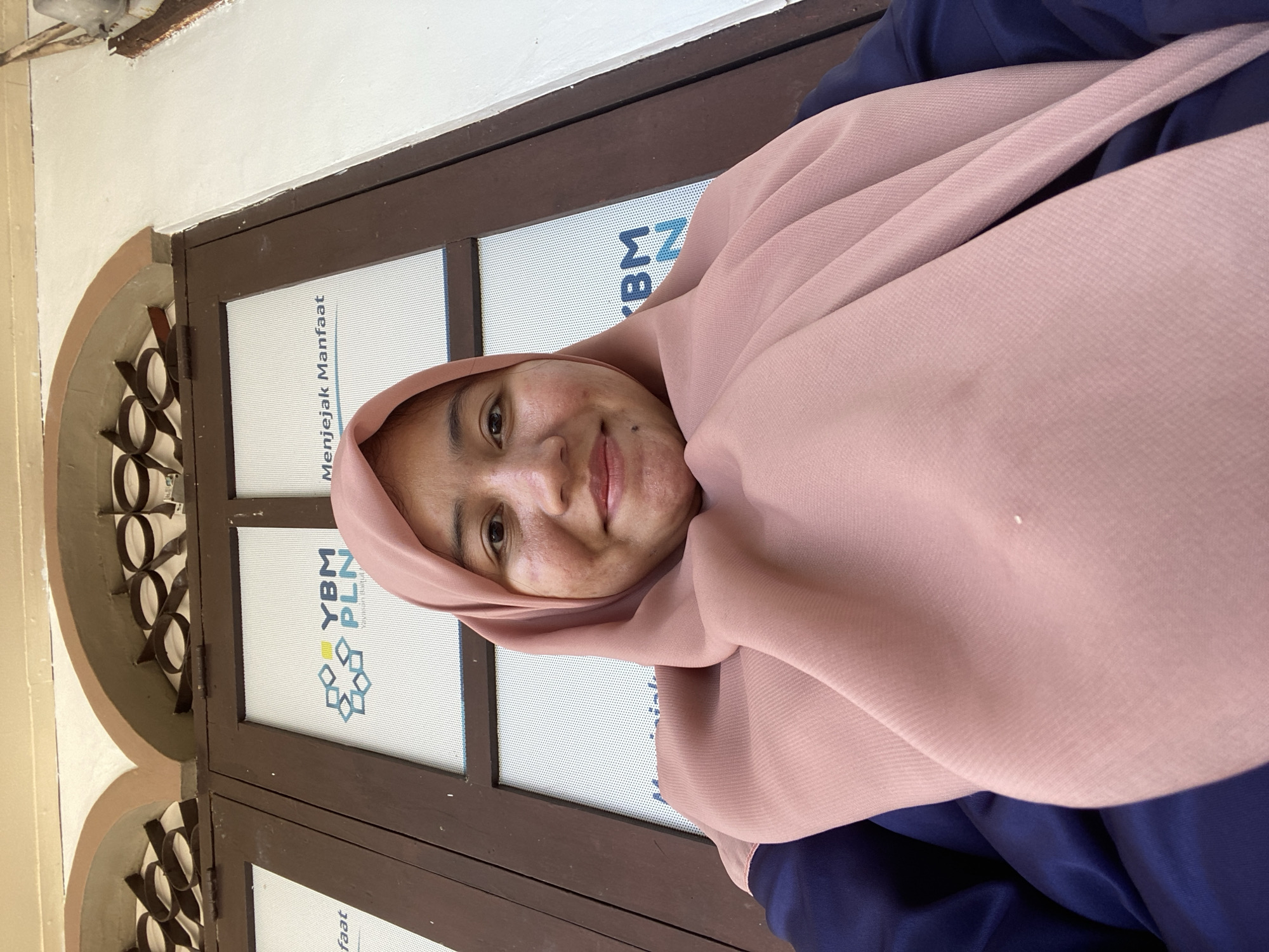 A woman wearing a pink hijab and a navy blue top is taking a selfie in front of a window. The window has dark brown frames and glass panels with white patterned film. On the film, there is text and a logo. The text reads "YBM PLN" and "Menjejak Manfaat". The logo is a stylized star shape with blue and yellow elements. The woman has fair skin and is smiling at the camera. The background suggests an outdoor setting with an architectural element above the window, possibly a building facade with arched openings and decorative metalwork. The overall impression is casual and outdoors, possibly in front of an office or public building.