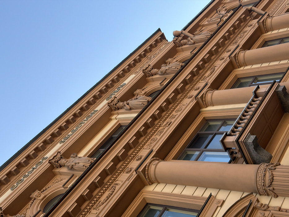 The image depicts an upward-angled view of the facade of the Cosmos Selection Hotel, located at Nevsky Prospect, 61, Saint Petersburg, Russia. The building features a multi-story, light brown/tan neoclassical architectural style against a clear blue sky.
The facade is extensively ornamented with relief carvings, cornices, and friezes. Large, fluted columns with decorative bases rise vertically, framing windows. On the lower visible level, a small balcony with a granite base and a balustrade extends from the facade. The windows are rectangular on the lower levels, transitioning to arched windows on the upper visible floor.
Along the upper portion of the building, a sign in relief lettering reads "COSMOS SELECTION ОТЕЛЬ" (Cosmos Selection Hotel). Directly below this sign, several ornate female figures, possibly caryatids, support architectural elements, positioned above the arched windows of the upper floor. These figures are uniformly colored with the building facade. A metal downspout is visible on the far right of the structure. No individuals, vehicles, or active interactions are present in the frame. The scene is static, illuminated by natural light that highlights the architectural details and casts subtle shadows.