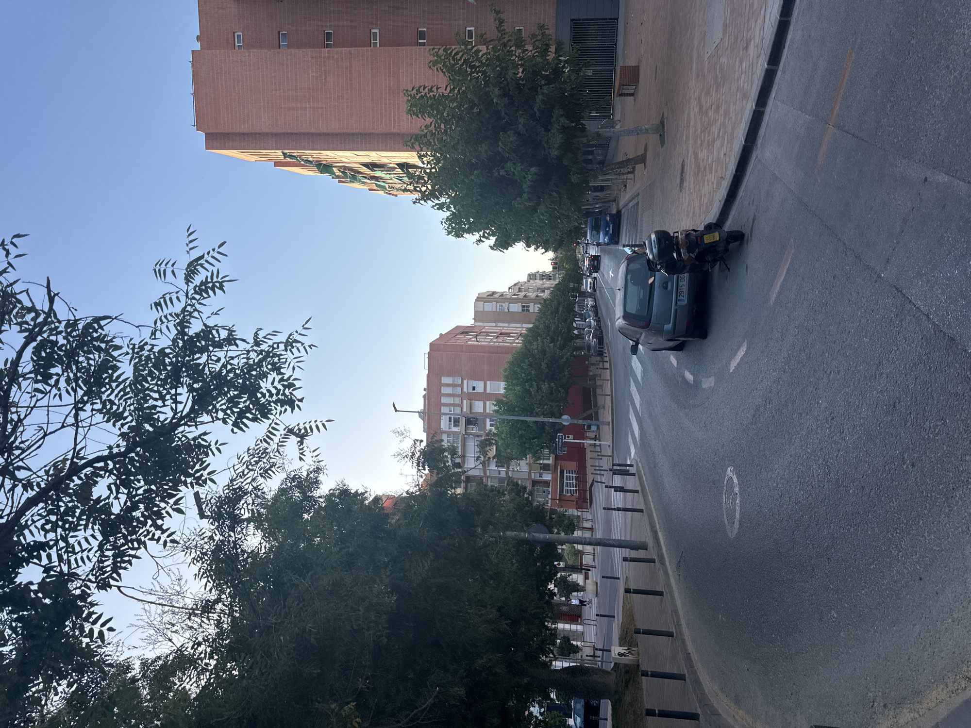 The image depicts a street scene in the city of Murcia, Spain. In the foreground, a curved asphalt road dominates the lower half of the frame, with a white dashed line marking its center and a solid white line bordering the right edge. A silver hatchback car is parked on the right side of the road, partially obscuring a black scooter parked behind it. The car's license plate reads "2915 DG". A circular manhole cover is visible on the road surface in the lower left quadrant.
To the right of the road, a paved sidewalk runs alongside a tall, reddish-brown brick building with multiple windows and green awnings. A metal fence and a bench are visible in front of this building. Further down the street, more multi-story buildings line the road, interspersed with trees.
To the left of the road, a sidewalk is lined with black bollards and mature trees with dark green foliage. Beyond the trees, other buildings and a crosswalk are visible. The sky is clear and blue, suggesting daylight. The overall impression is an urban street during the daytime.