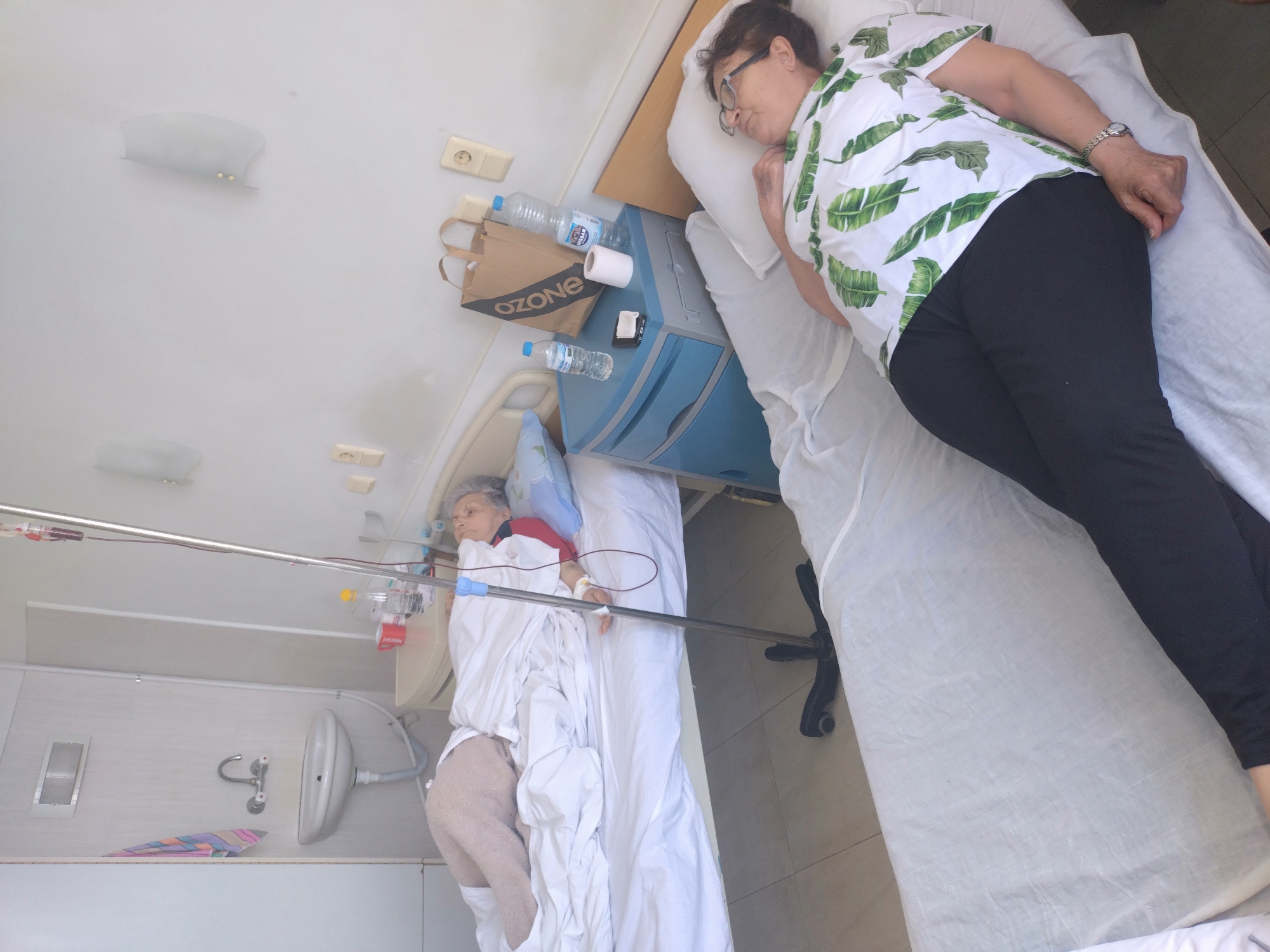 This media file captures an indoor scene within what appears to be a hospital or clinic room in Stara Zagora, Bulgaria. The image is oriented vertically, showing the room from an overhead perspective, with subjects lying horizontally.
In the foreground, an older woman is seen sleeping peacefully on a bed. She is wearing a white short-sleeved shirt with a green tropical leaf pattern and black pants, along with eyeglasses. Her head rests on a white pillow.
Adjacent to her, separated by a blue and grey bedside cabinet, another woman, presumably a patient, lies on a second hospital bed. She has light-colored hair and is partially covered by white sheets. An IV drip, connected to a metal pole with tubing and a small red medical device, leads to her arm, indicating she is receiving treatment.
The bedside cabinet holds several items, including two plastic water bottles (one with "S.PELLEGRINO" visible), a roll of toilet paper, and a brown paper bag prominently labeled "OZONE". In the background, a small white sink with a faucet is mounted on the wall. The room also features wall-mounted lights and electrical outlets. The lighting suggests it is daytime. The overall atmosphere appears calm, centered around rest and medical care.