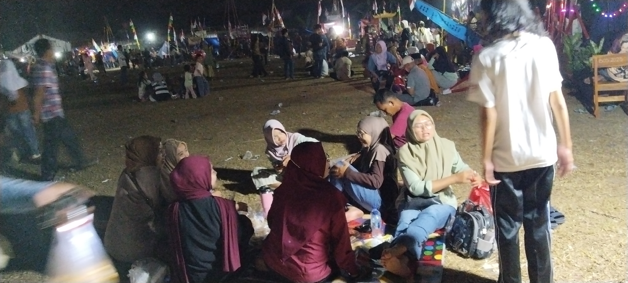 A nighttime outdoor gathering is taking place on a dirt or gravel surface. In the foreground, several individuals are seated on the ground, some on mats or blankets. A group of women in hijabs are interacting. One woman in a light green hijab is sitting with her legs extended, holding a red plastic bag. Another woman to her left, also in a hijab, is looking down. A woman in a dark red hijab is facing away from the camera, and another woman in a brown hijab is seated to her left. To the left, partially visible, is a person wearing a white shirt and black pants with white stripes. In the background, a crowd of people are standing and milling about. There are various flags on poles, some with text and graphics, and several tents or canopies are visible. The lighting is dim, suggesting it is nighttime, with artificial lights illuminating the scene from various points. Some people are wearing casual clothing. The overall impression is of a community event or festival in Sido Rahayu, Indonesia.