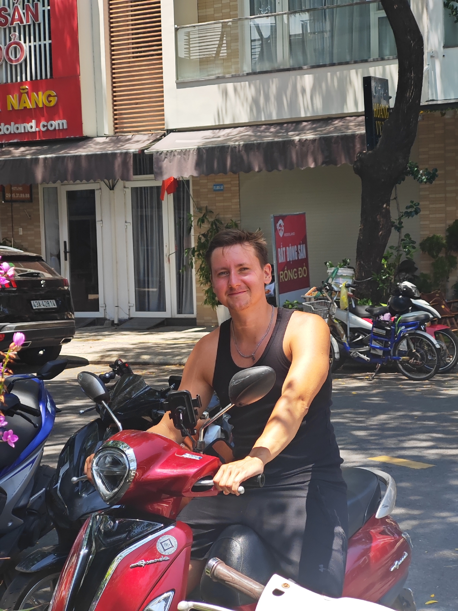 A man is seated on a red scooter in a city setting. He is wearing a black tank top and black shorts, with a silver chain necklace. He is smiling at the camera and his right hand is on the scooter's handlebar. The scooter is parked on a paved road. In the background, there are several other scooters and a black SUV. A building with signs in Vietnamese is visible behind the scooters. The scene appears to be in the city of Đà Nẵng, Vietnam. The man's posture and the presence of scooters suggest a typical street scene in Vietnam.
